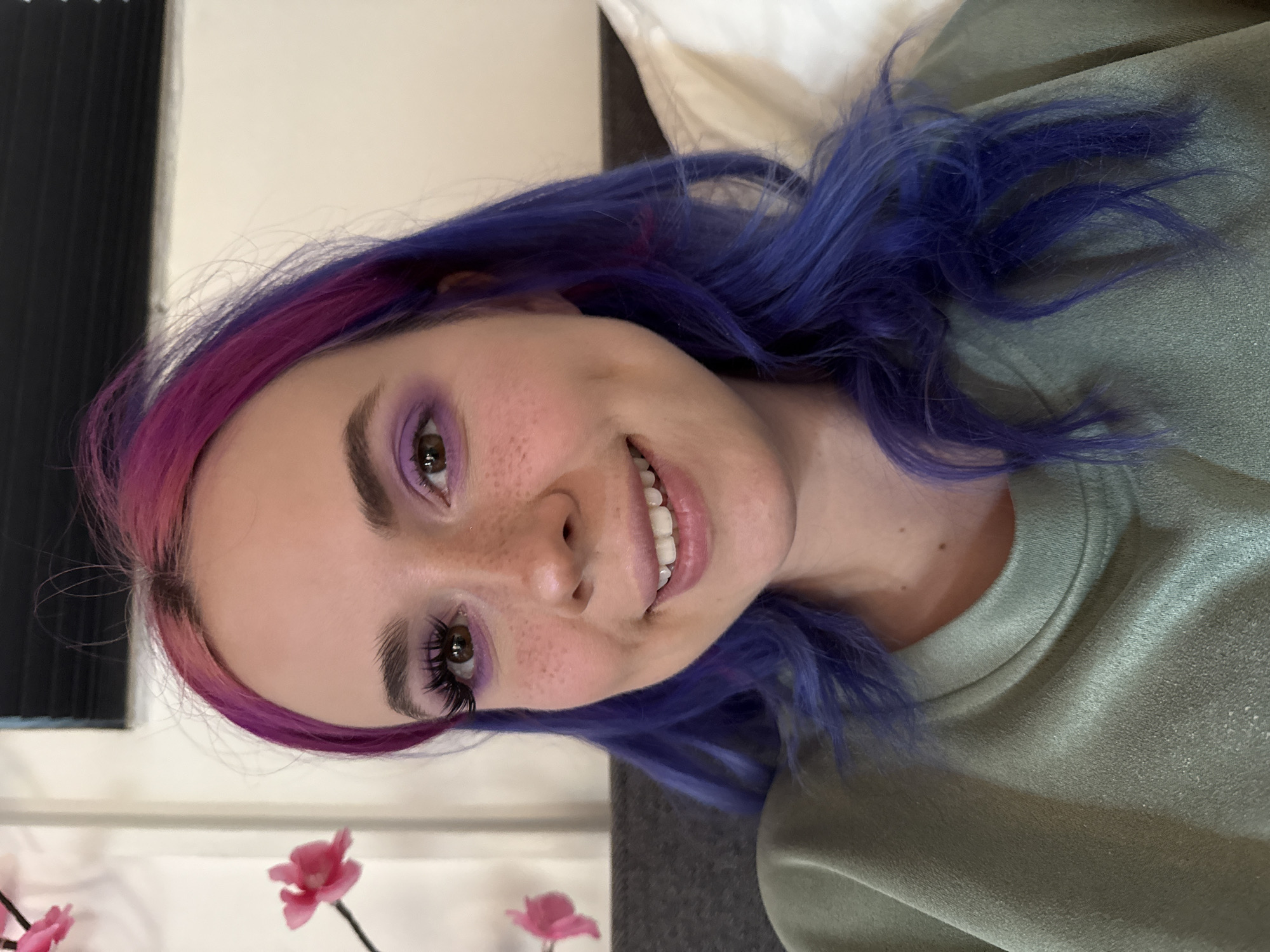 A medium close-up shot shows a woman with dyed hair lying on her side. The top of her hair is pink and transitions into purple on the sides. She has light skin, freckles, and brown eyes with purple eyeshadow. Her eyes are open and she is smiling, revealing her teeth. She wears a green shirt, and her hair is resting on the shirt. In the background, to the left, there is a window with vertical blinds and, to the right, a white pillow is visible. There are pink flowers out of focus at the bottom of the image.