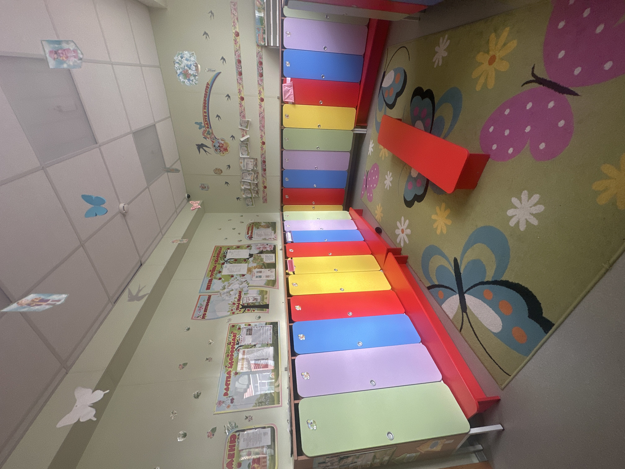 The image depicts a brightly colored room, likely a kindergarten or preschool classroom in Novoaltaysk, Russia. The room features a row of lockers in various pastel colors, including green, purple, blue, red, and yellow. A red bench runs along the front of the lockers. The floor is covered with a green carpet adorned with large, colorful butterfly and flower motifs. The walls are painted a light green and decorated with various posters, charts, and cut-out paper decorations, including butterflies and birds. Some of the posters appear to be educational, with text in Russian. The ceiling is white and appears to be a drop ceiling with fluorescent lighting. The overall impression is of a cheerful and child-friendly environment. There are no people visible in the image.