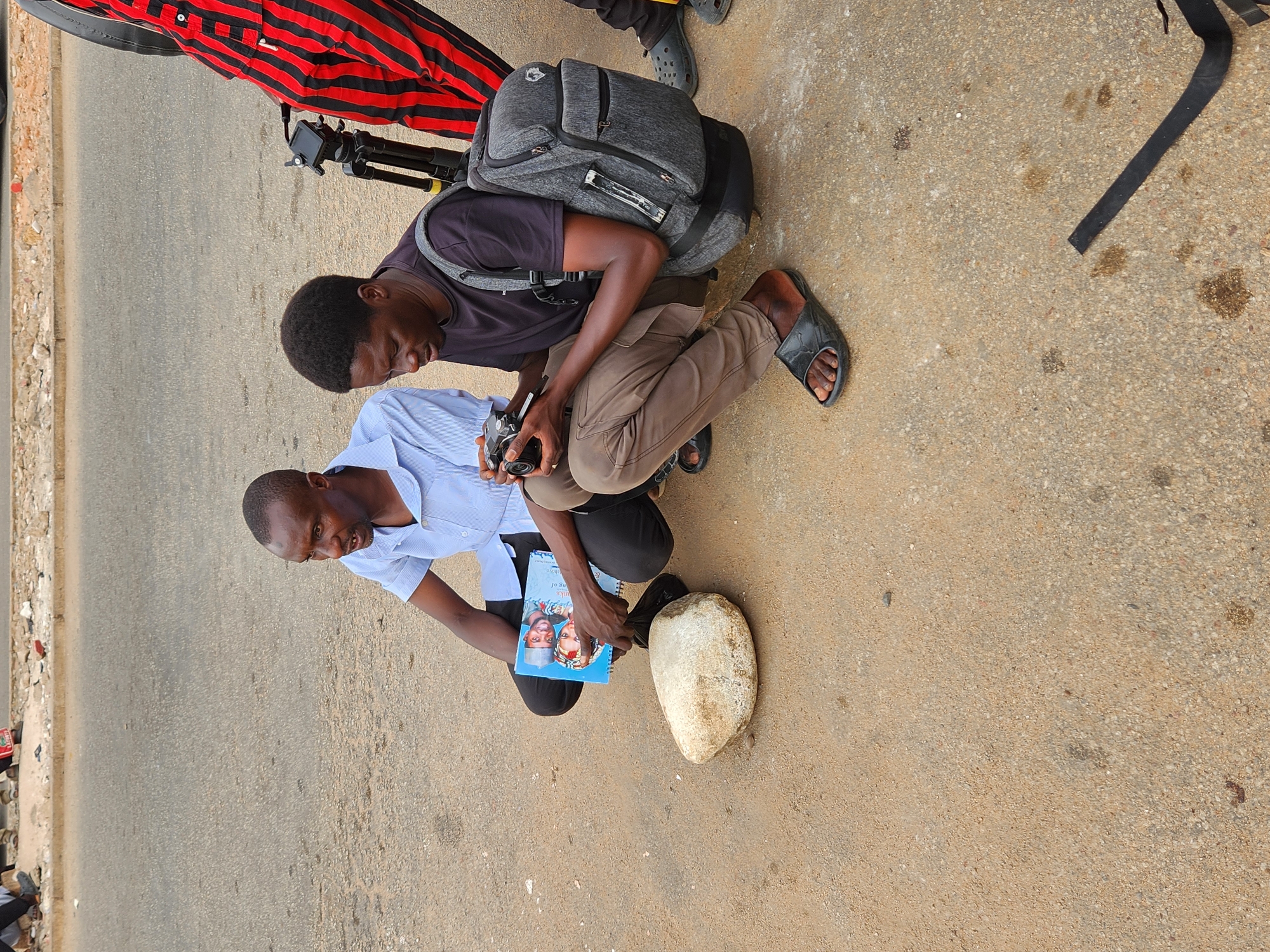 Two men are kneeling on a paved road. The man on the left wears a light blue shirt and black pants. He holds a blue book with a photo of a couple on the cover, resting it on a light-colored, elongated rock or object. The man on the right, wearing a dark purple shirt and tan pants, operates a camera, focusing on the rock/object. He has a gray backpack. A black and red striped garment or object lies near them, as does a camera tripod. The location is presumably Jos, Nigeria, given external context. The road surface is worn, with scattered debris.