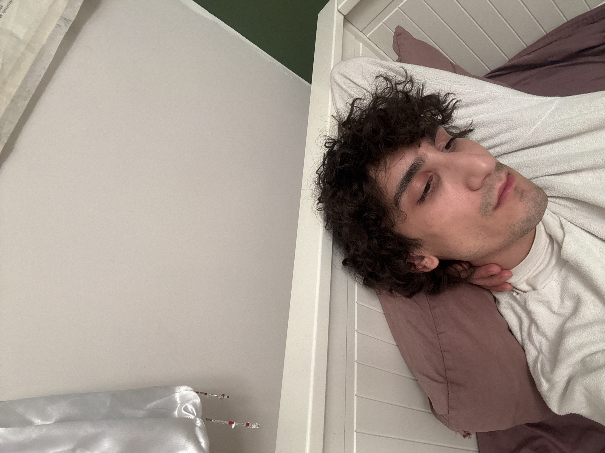 The image shows a young man sleeping or resting in a bedroom in Moscow, Russia. He is positioned on his right side, with his head on a mauve-colored pillow, and his eyes are closed or partially closed. He has dark, curly hair and some stubble on his face. He wears a white, possibly textured, long-sleeved shirt or sweater. One hand is partially visible near his chin, appearing to support his head.
The setting is a bedroom, featuring a white slatted headboard behind the man. To the left, a plain white wall is visible, with a section of dark green paint appearing in the upper left corner, possibly an accent wall. In the bottom left of the frame, there is a piece of white satin-like fabric and two decorative items resembling skewers or sticks with red beads on their ends. The lighting suggests an indoor environment, creating a calm and restful atmosphere. No visible text is present in the image.
