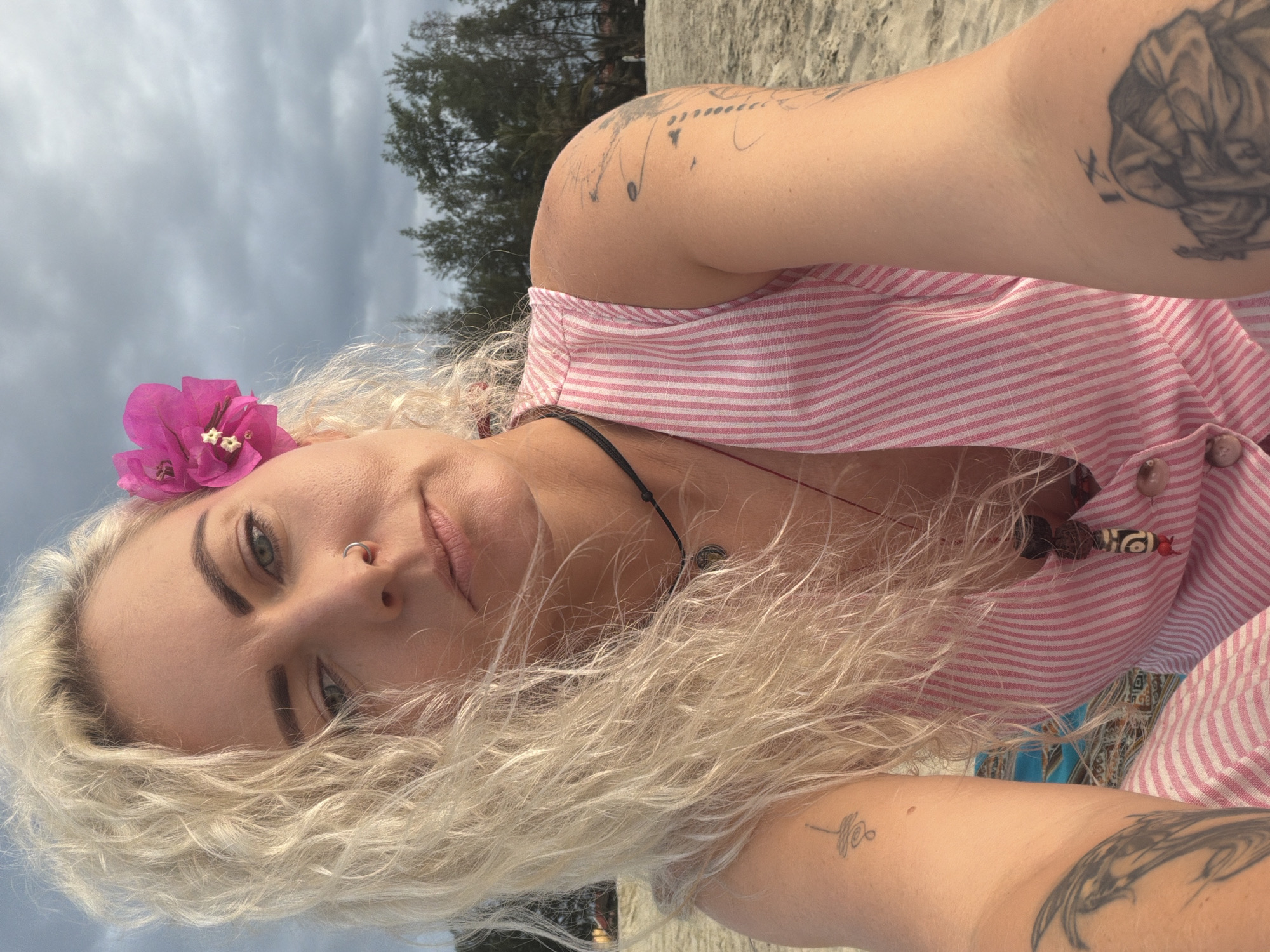 A woman with blonde, curly hair and a pink flower tucked behind her ear takes a selfie on a beach. She is wearing a pink and white striped sleeveless top and several necklaces. She has tattoos on her left arm and shoulder. The background shows a cloudy sky, trees, and the sandy beach. The scene is located on An Bang Beach in Hoi An, Vietnam.