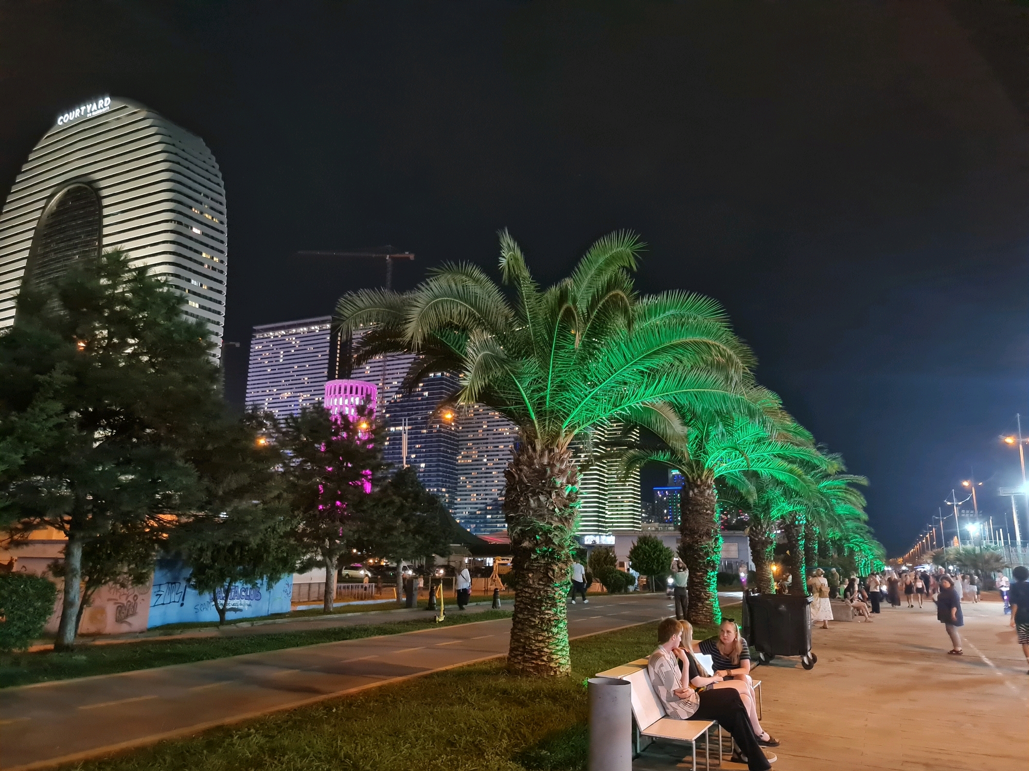 The image depicts a night scene in Batumi, Georgia, likely on the Batumi Boulevard, a prominent seaside promenade. The foreground features a paved walkway and a grassy area with several palm trees, some of which are illuminated with vibrant green light. Two individuals are seated on a white bench, engaged in conversation. Behind them, a line of large, dark trash bins is visible. Further into the background, a variety of buildings are illuminated against the dark sky. The most prominent is a distinctive white, curved skyscraper with horizontal banding, identified by a "COURTYARD BY MARRIOTT" sign at its apex. Adjacent to it are other high-rise buildings with illuminated windows. A pink-lit structure, possibly a tower or decorative element, is also visible. A road or pathway runs parallel to the boulevard, with people strolling and some parked vehicles. The overall atmosphere suggests a lively urban environment at night, with architectural landmarks and recreational spaces.