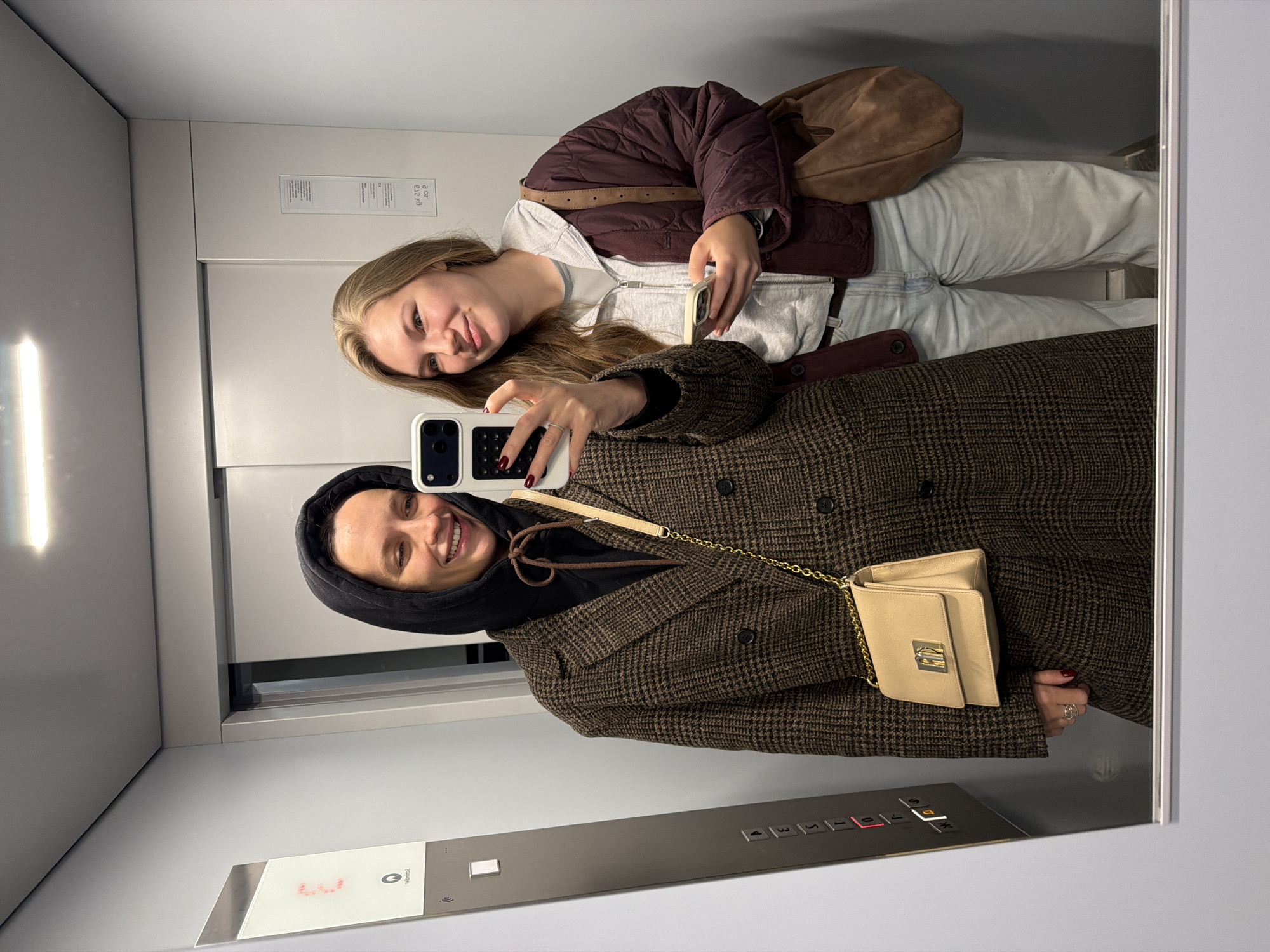 Two women take a selfie in an elevator with a mirrored wall. One woman wears a brown plaid coat and a black hoodie. She holds a white smartphone in her right hand. The second woman wears a burgundy quilted jacket, a light grey zippered sweatshirt, and light blue jeans. She carries a brown leather purse over her right shoulder. The elevator has a stainless steel control panel with numbered buttons. The photo was probably taken on the ground floor, as the display shows "G." The location is Vozdovac Urban Municipality, Serbia.