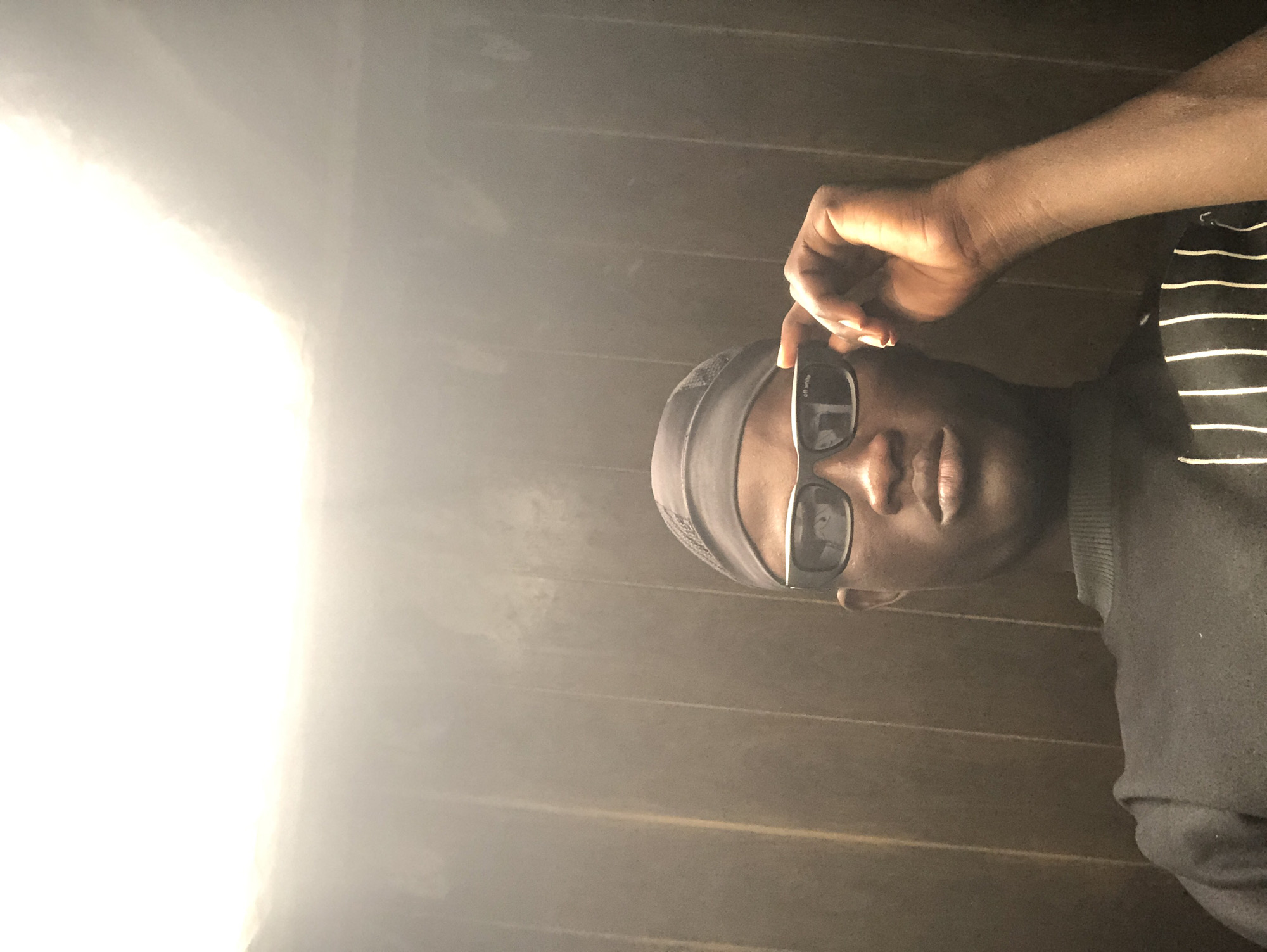 In this low-angle selfie, a young Black person is positioned centrally, with their head tilted slightly down. They are wearing a dark durag and dark-rimmed sunglasses with a white "Off-White" logo visible on the left lens. Their right hand is raised, with the index finger and thumb lightly touching the temple of the sunglasses. They are wearing a dark, high-necked sweater with white vertical stripes across the chest. The background consists of dark, vertical wood paneling. A bright light source is positioned above and to the left of the frame, casting a strong glow. The overall lighting is dim, except for the direct illumination from the top. There are no visible indicators of the specific location in Ado, Nigeria.