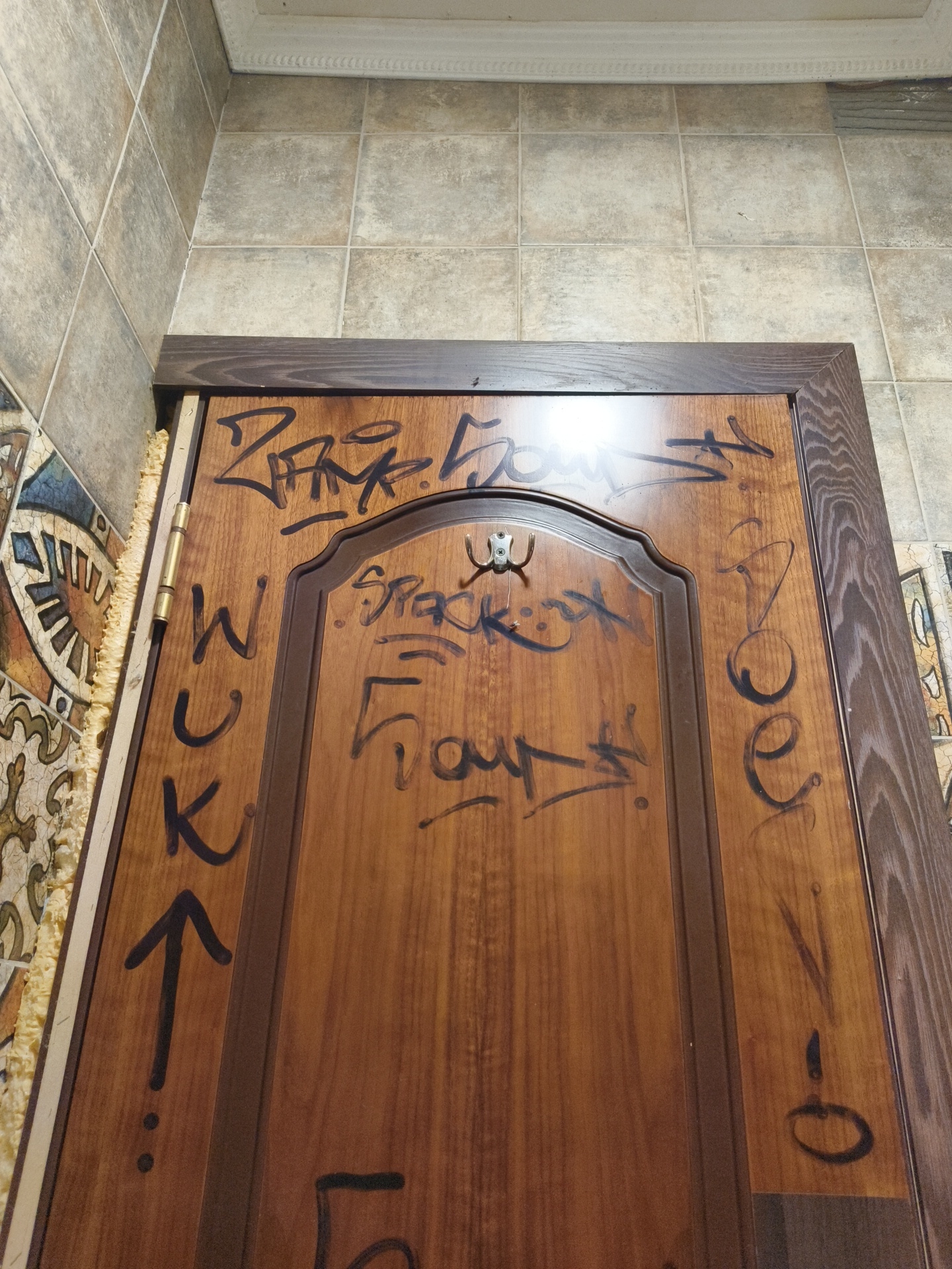 The image shows a wooden door with black graffiti spray-painted on it. The graffiti includes stylized letters and symbols. The door is framed by a darker wooden trim and is set against a tiled wall. A metal hook is visible on the door. The overall scene suggests a public or semi-public space that has been vandalized. The location is not specified in the image.