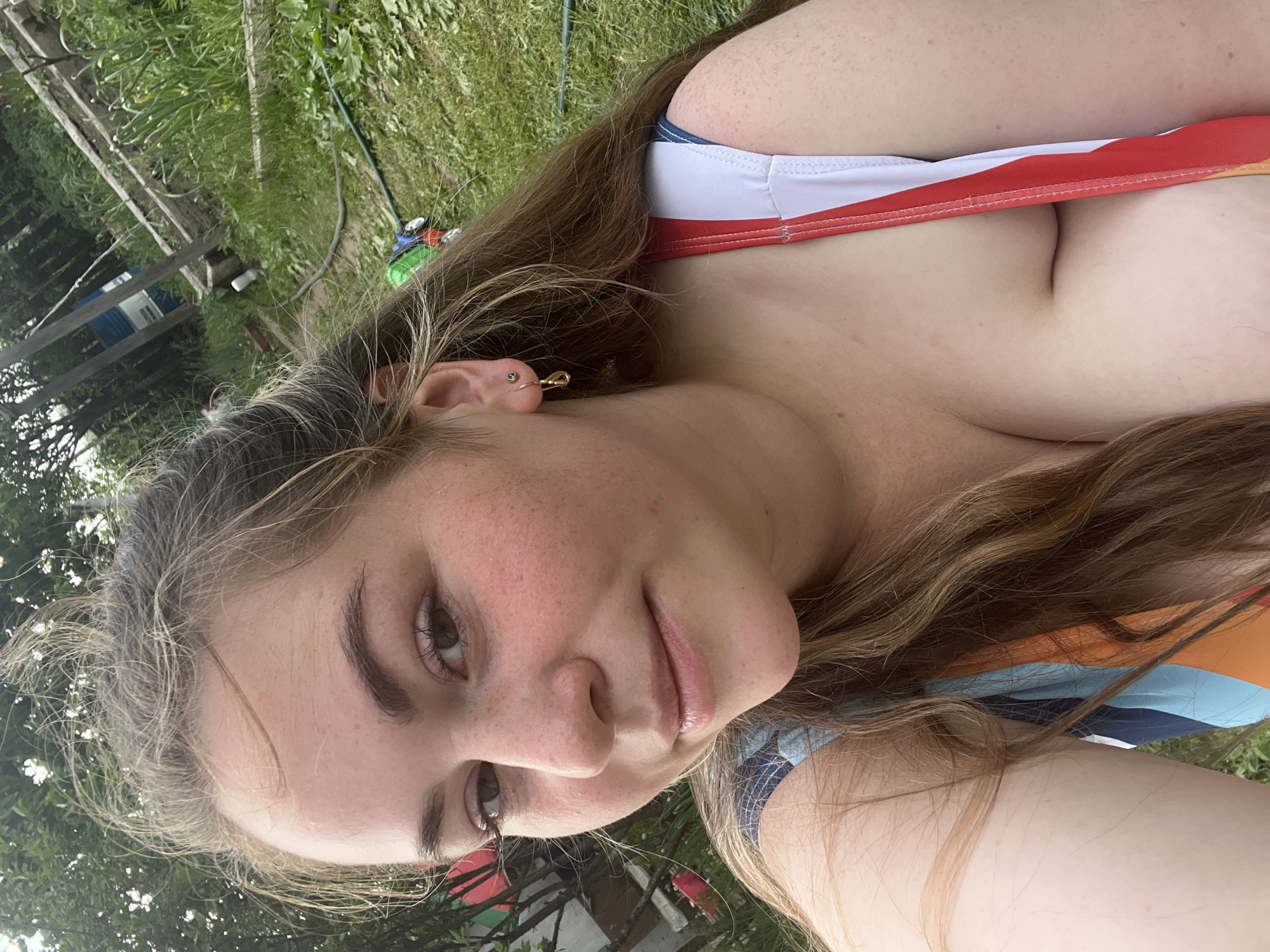 A fair-skinned woman is photographed outdoors, likely in a grassy area. She has brown hair and visible freckles on her face. She is wearing a swimsuit with a color scheme of white, red, and blue. Her hair is partially pulled back. Her left ear is visible, with what appears to be multiple earrings. The background includes greenery, suggesting a park or garden setting. It is not possible to determine the location as Chaikovsky, Russia from the image.