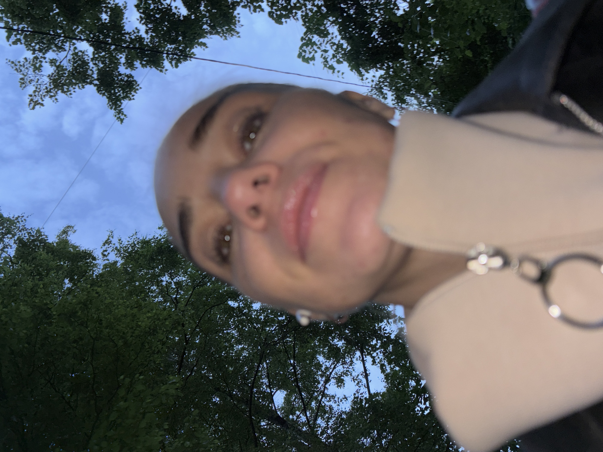 The original image is a close-up, low-angle selfie of a person's face, predominantly out of focus, with trees and a cloudy sky in the background. The subject's head is tilted upwards, and their face is partially obscured by the camera's proximity. They appear to be wearing a light-colored jacket with a visible zipper pull featuring a silver charm. The overall lighting suggests it may be dusk or early morning. The image's composition and lack of identifying landmarks make it impossible to determine the specific location within a city, let alone "Unknown, Russia."