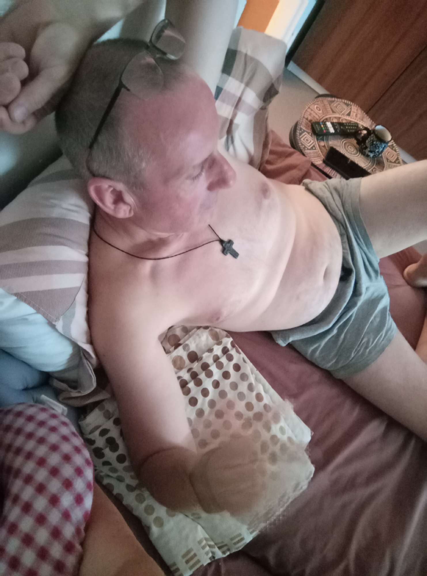 The image shows a man lying in bed. He is wearing grey shorts and a necklace with a cross pendant. Glasses are resting on his head. The bed appears to be in a bedroom, with pillows and blankets visible. A bedside table with remote controls and a cup is also in the frame. The lighting suggests it is daytime. There are no specific details in the image that can indicate the location in the city of Brno, Czechia.