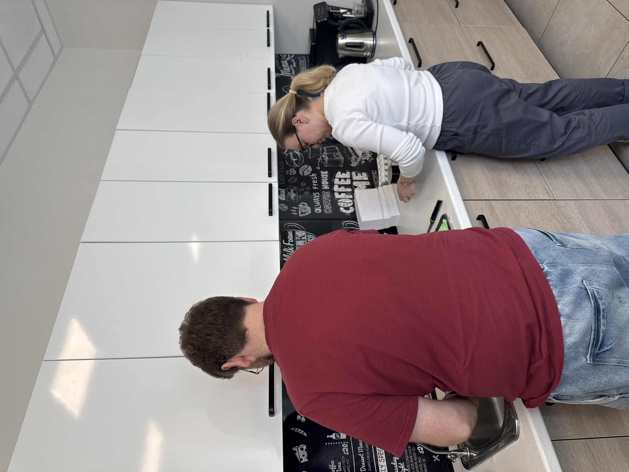 This image captures two individuals at a modern coffee station or breakroom, likely within an office or commercial setting, located in Moscow, Russia. The scene features clean white overhead cabinets, a light-colored countertop, and a black backsplash decorated with white chalk-style lettering and illustrations.
On the left side of the upright image, a man with dark, short hair and a beard, wearing a red t-shirt and blue jeans, is leaning forward over the counter. He is positioned near a stainless steel sink with a faucet, suggesting he may be using the water or cleaning something.
Further along the counter to the right, a woman with blonde hair tied in a ponytail, wearing a white long-sleeved top and dark pants, is also leaning forward. She is situated near a stack of white paper cups or napkins and what appears to be a coffee machine and an electric kettle. Both individuals appear focused on tasks at the counter, possibly preparing beverages or engaging with the coffee station equipment.
The black backsplash prominently displays various coffee-related texts and menu items. Visible phrases include "Menu", "ALWAYS FRESH", "Best coffee", "COFFEE HOUSE", "COFFEE ME", and "PREMIUM", accompanied by small graphic icons. A more detailed menu section is also visible, listing "Coffee Menu", "ALWAYS FRESH ALWAY HOT", "$20:", "Dessert Menu", and snippets such as "LY SPEC", "Tuesday", "night", and what appears to be Latin-like placeholder text: "MOD CONSEQUA", "SAUTE IRURE DOLOR", "REPREHE NDERIT DO", "SMOD MCO", "ABORIS SE", along with a "$2" price. The overall environment is bright and minimalist.