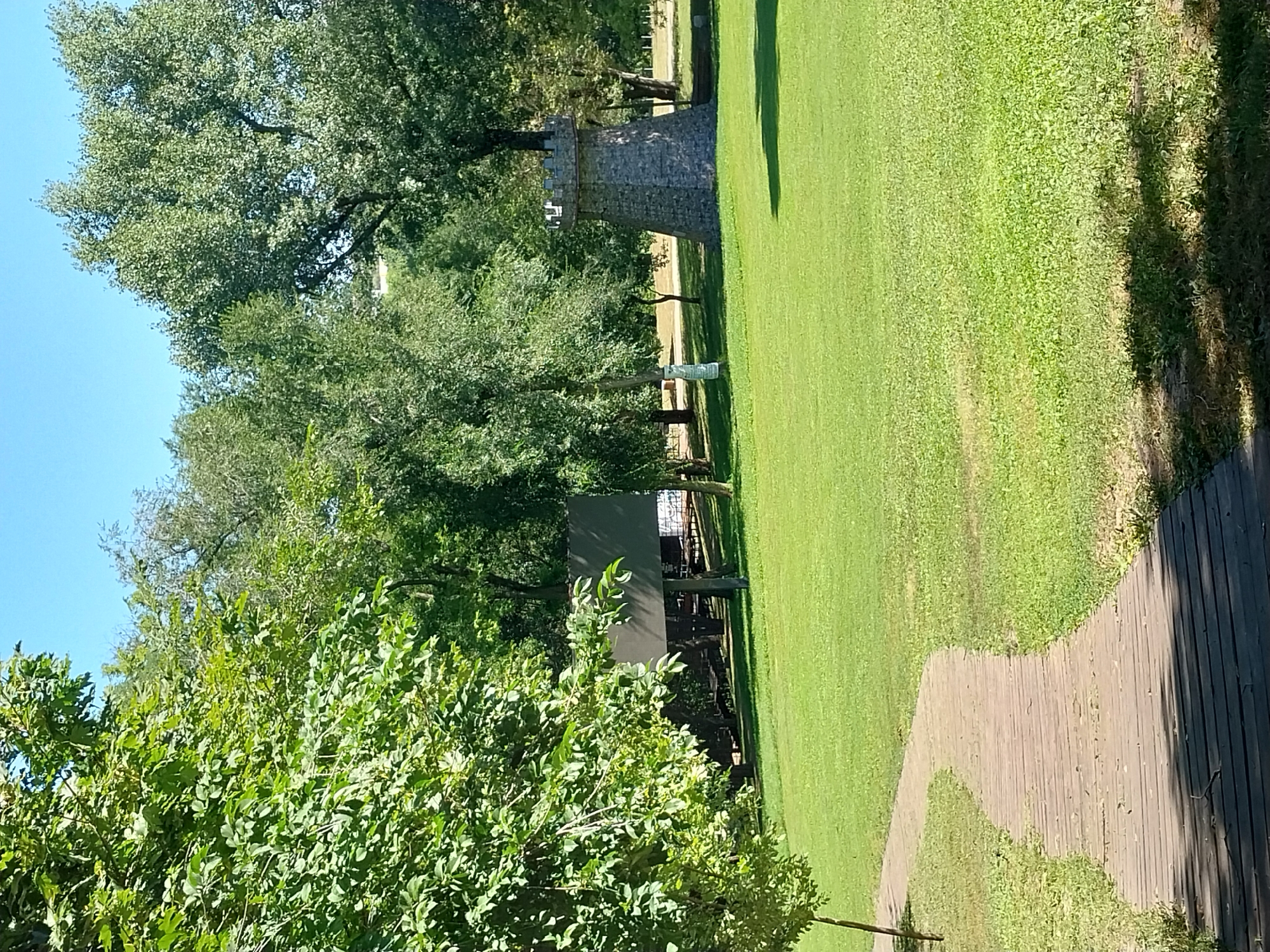 The image captures a sunlit scene in a park setting. A winding wooden boardwalk leads from the lower left corner into a well-maintained grassy area. In the middle ground, a large, dark, rectangular structure, possibly a billboard or informational display, is visible behind some trees. Further to the right, a stone tower, reminiscent of a small castle turret with crenellations, stands on the grass. The background is filled with lush green trees under a clear blue sky. The overall impression is one of a recreational area, likely a park within the city of Dnipro, Ukraine. There are no people or animals visible in the frame.