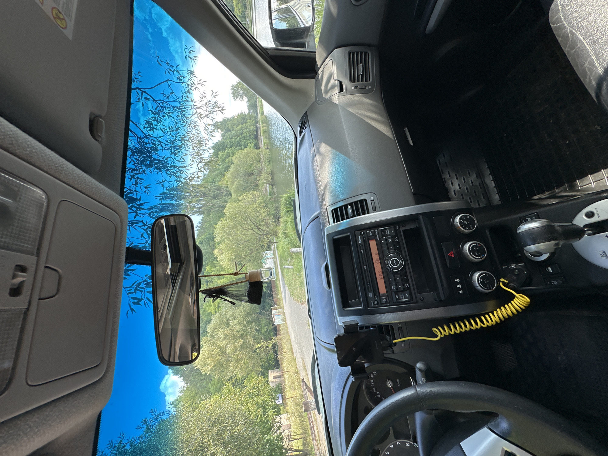 The image shows the interior of a car parked on a dirt road with a lake and trees in the background. The car's dashboard is visible, featuring a radio, air conditioning vents, a steering wheel, and a gear shifter. A yellow coiled cable is plugged into the dashboard. The car is likely in Makiv, Ukraine, given the context. The environment appears to be rural, with natural scenery and a body of water. There are no people visible in the image.