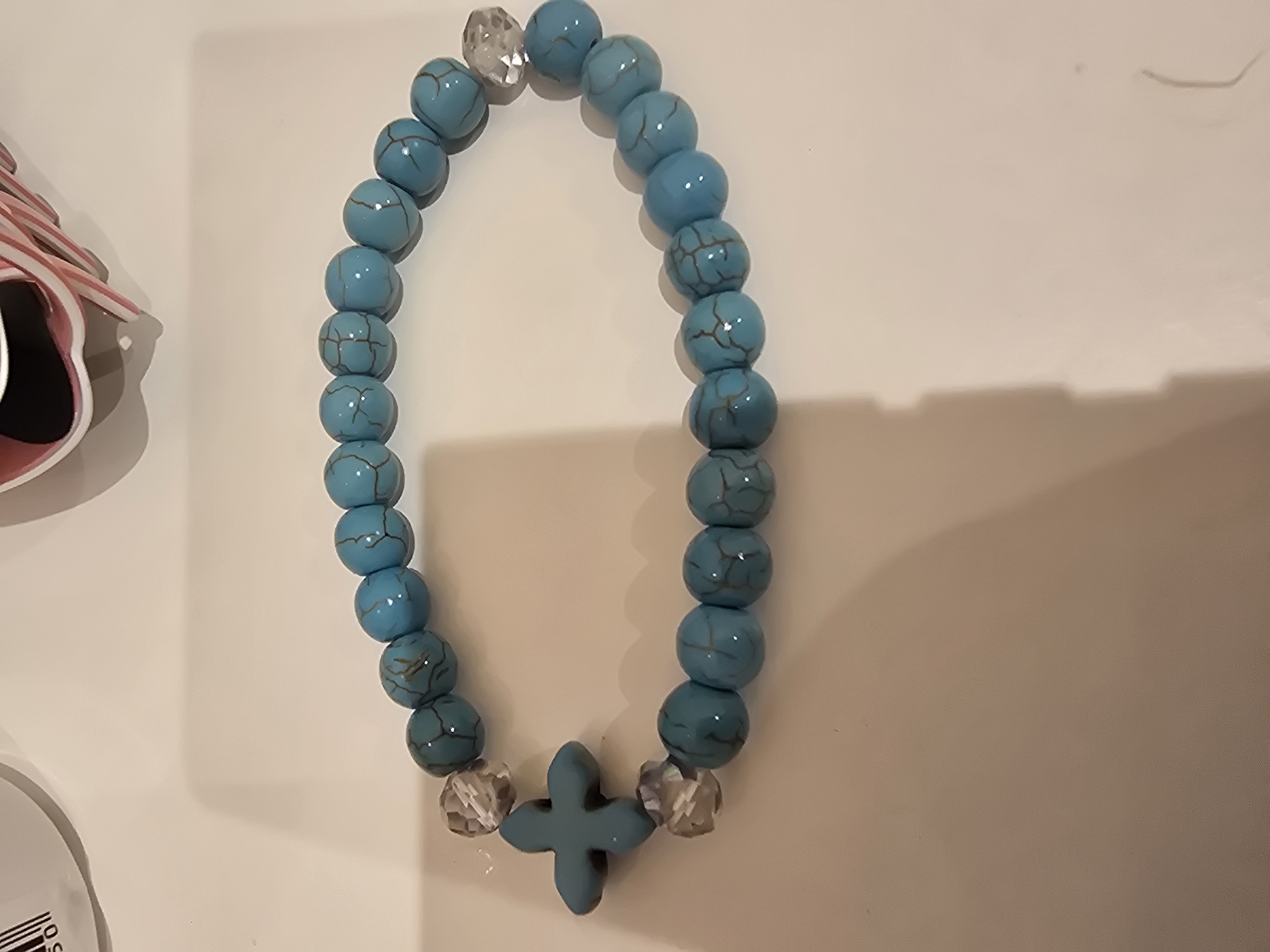 A close-up, overhead shot shows a turquoise beaded bracelet with a cross charm. The bracelet is laid out on a light-colored, flat surface, likely a table or countertop. The beads are round and uniform in size, with a natural, veined pattern in a darker shade of turquoise. Between some of the turquoise beads, there are clear, faceted spacer beads. The charm at the center of the bracelet is a small, turquoise cross. The lighting casts shadows to the right of the bracelet, suggesting the light source is from the left. There are faint out-of-focus objects in the background, one appearing to be a white cylindrical container with some black markings, and another with pink and white stripes. The overall impression is that of a handmade piece of jewelry.