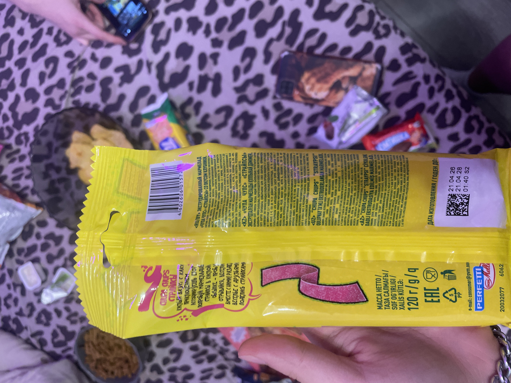 The image displays a close-up of a yellow package of Chupa Chups candy, held by a person's hand. The package features prominent branding and nutritional information. The background is blurred, but suggests a domestic setting with a leopard-print fabric and other objects. There is no visible indication of the city of Zmeinyi, Russia, in the image. The lighting appears to be natural, possibly from a window.