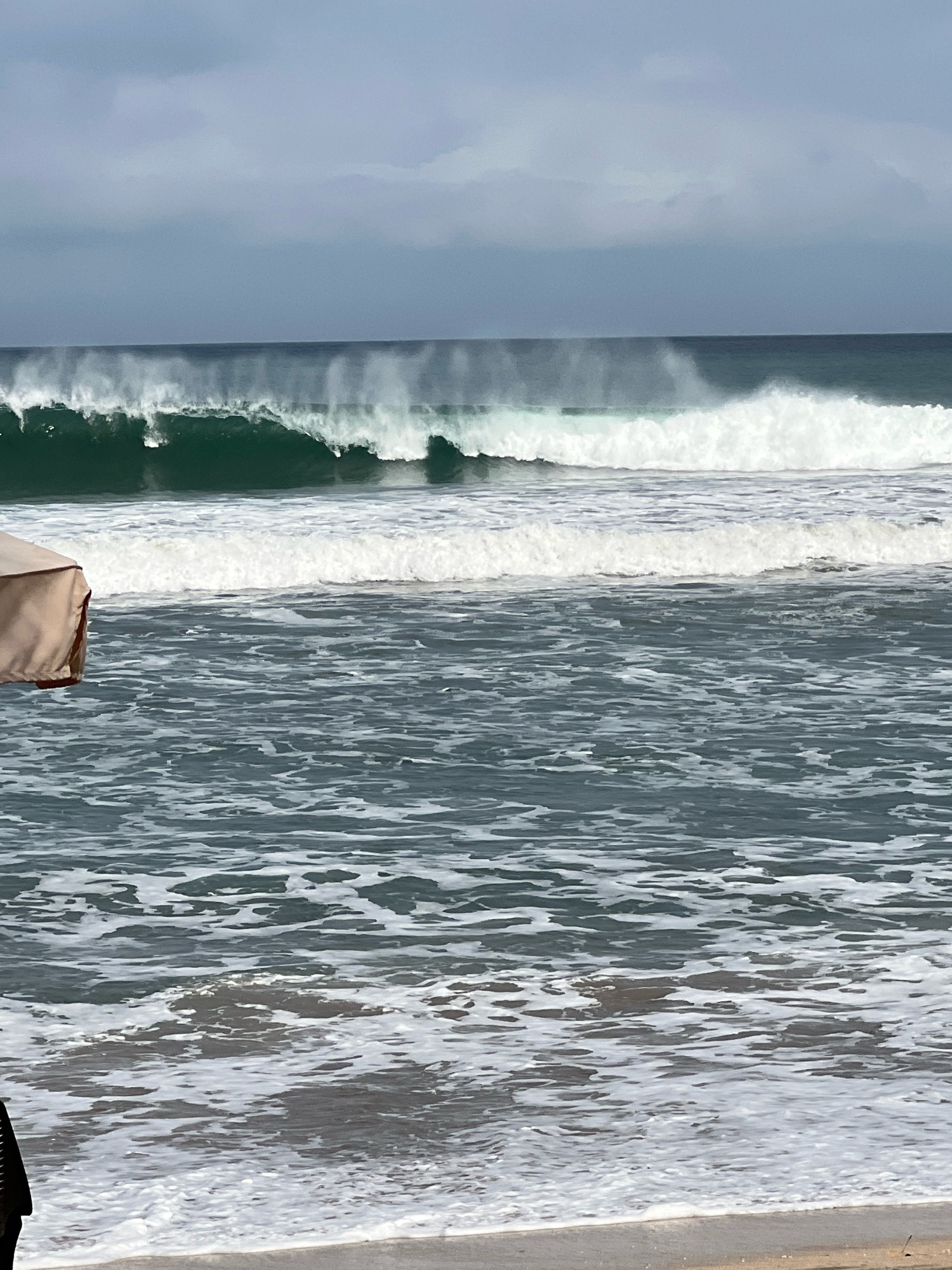 An ocean scene in Kuta, Indonesia, features large waves breaking on the shore under a cloudy sky. The waves are dark green and capped with white foam, with spray misting off the crests. In the foreground, a portion of a tan beach umbrella is visible on the left side of the frame, and a dark object, possibly a person's leg or clothing, is at the bottom left edge. The water is choppy with foam and whitecaps reaching the sandy beach. The overall impression is of a dynamic and somewhat overcast day at the beach.