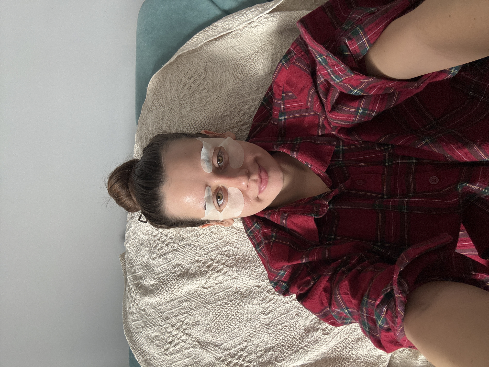 A woman lies on a textured, light-colored blanket on a turquoise surface (likely a sofa or bed). She is wearing a red plaid shirt. She has eye patches under both eyes. Her hair is styled in a bun. A light-colored wall is visible in the background. The location cannot be determined from the image.
