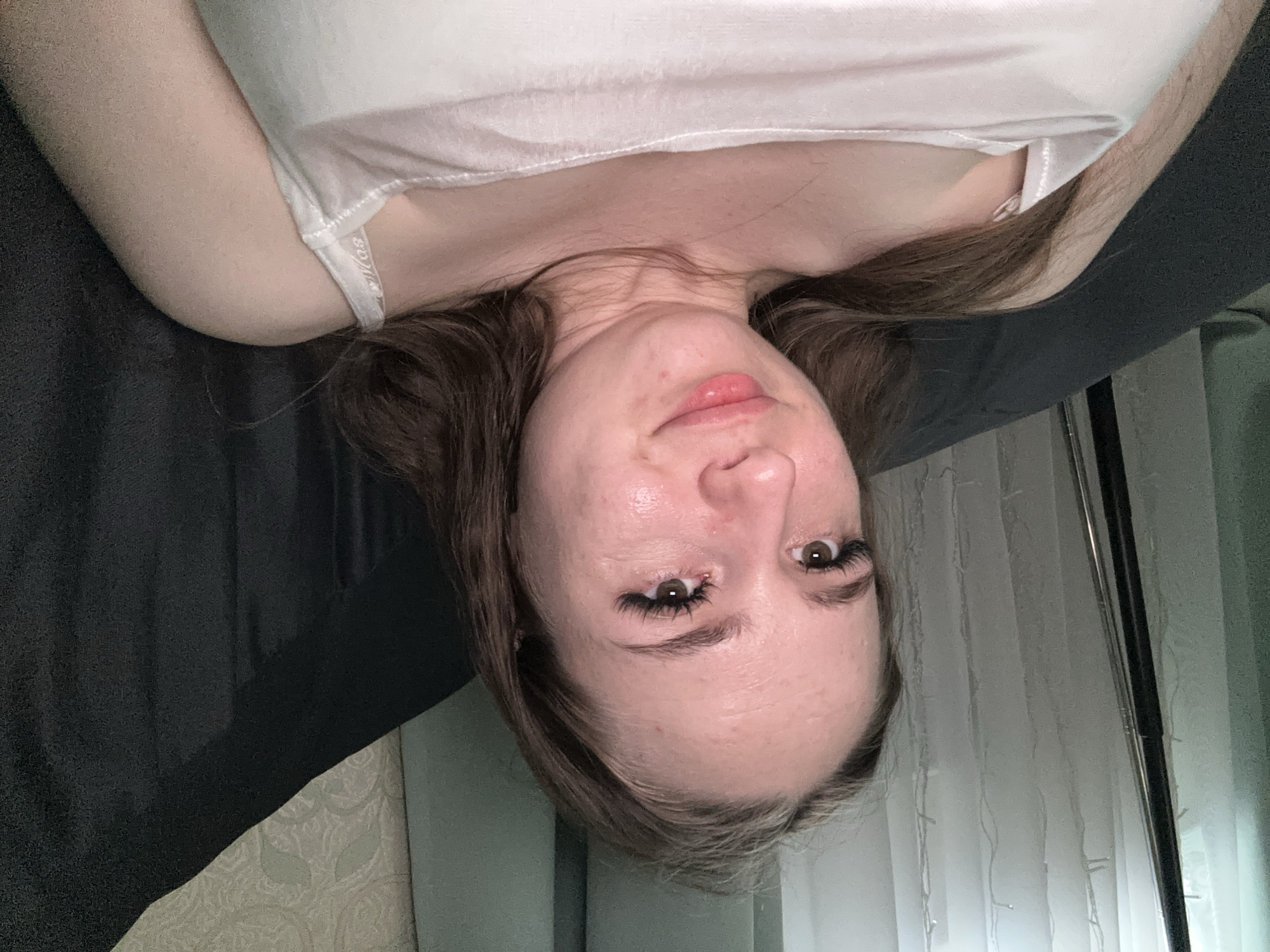 The media file shows a close-up, upside-down selfie of a young woman lying down. She is wearing a white tank top, and her brown hair is spread out. Her face is the primary focus, with her eyes open and looking towards the camera. She has noticeable eyelash extensions. Her lips are slightly parted, and her skin appears to have a subtle glow, possibly from lighting or skincare products. The background is dark on the left side, suggesting a shadowed area, and transitions to lighter textures and vertical blinds with faint lights on the right. The lighting seems to be coming from the right side, illuminating her face. The overall mood appears relaxed and casual. No text is visible in the image.