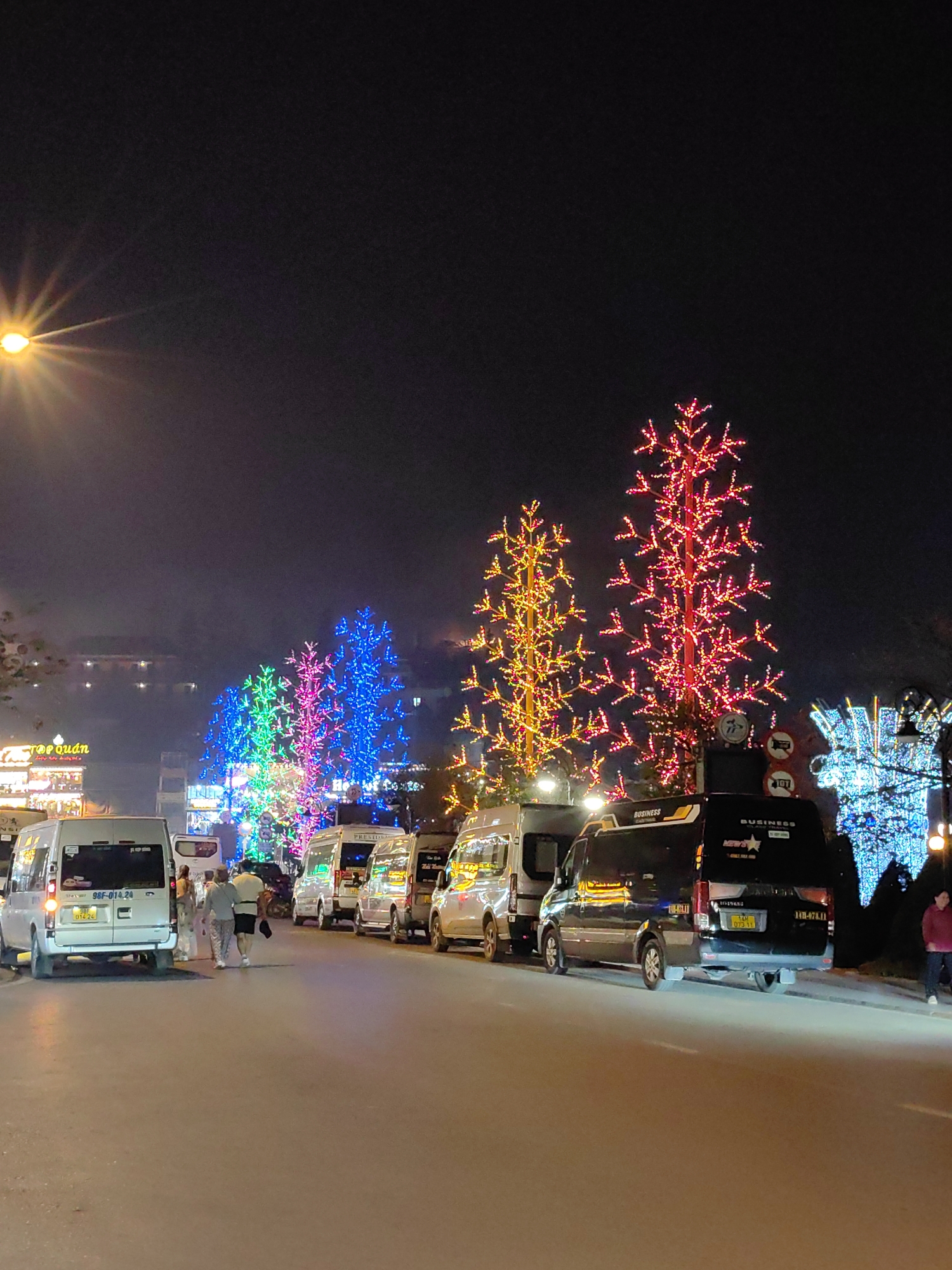 This image depicts a nighttime street scene in Sa Pa, Vietnam. A row of vehicles, predominantly white and black passenger vans or minibuses, are parked or stationary along the right side of a paved road. Many of these vans feature "BUSINESS" text on their sides or rear doors; one visible black van has license plate 19H-078.11 and another white van has license plate 98F-014.24.
The street is illuminated by various light sources. A bright yellow streetlight with a starburst effect is visible in the upper left corner. In the background, a series of decorative, artificial trees illuminated with vibrant LED lights line the street. These installations display a spectrum of colors including red, yellow, green, blue, and purple. Building facades with illuminated signs, one partially reading "TOP QUAN", are visible further back on the left.
Several pedestrians are present. Two individuals, a man and a woman, are walking away from the camera towards the middle-ground, positioned between parked vehicles. Another person in a red jacket is visible on the far right near the curb. Circular road signs are partially visible between the illuminated trees. The overall environment is an urban setting, busy with artificial light and activity at night.