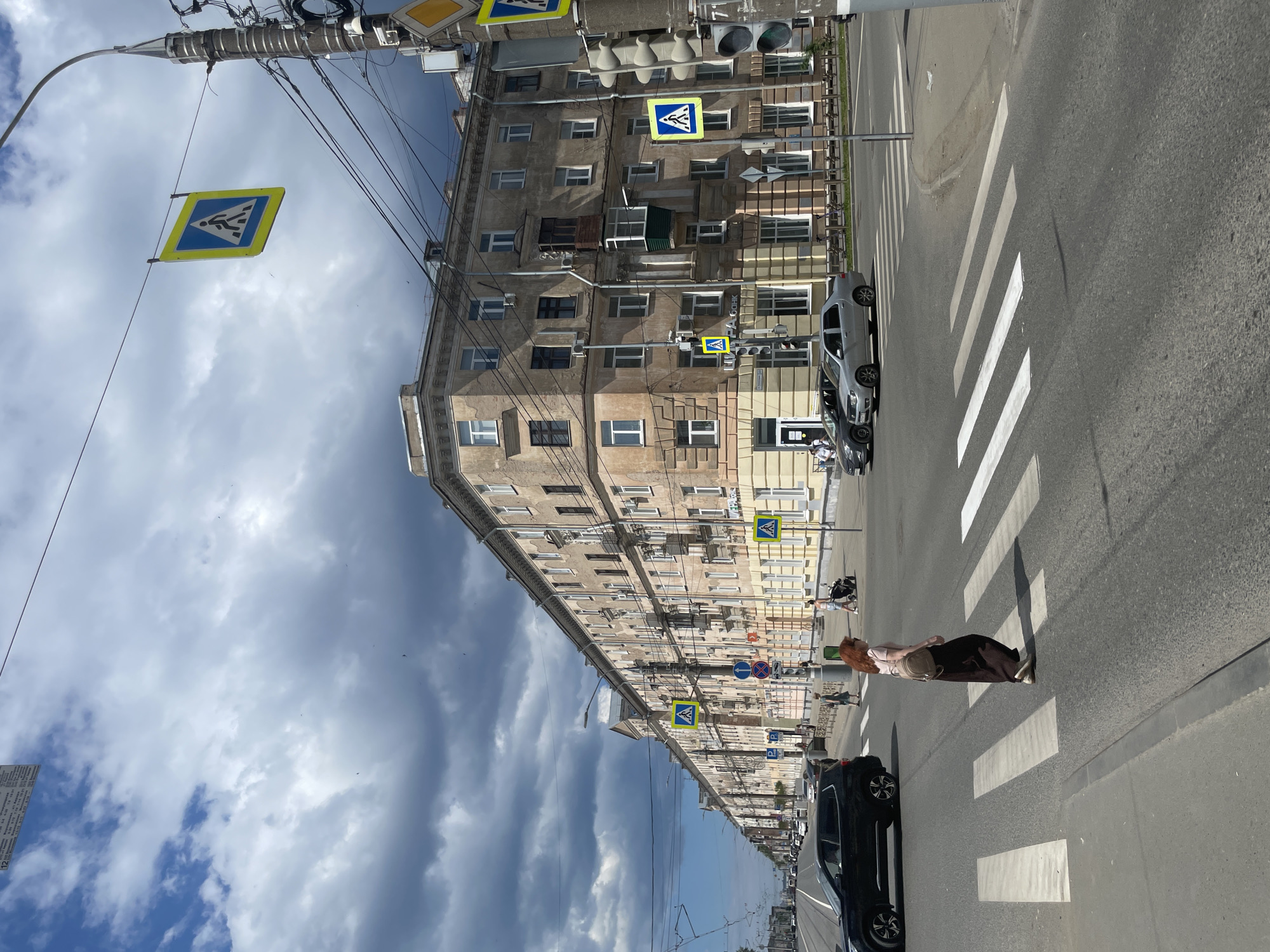 The image shows a street scene in Izhevsk, Russia, on a cloudy day. A pedestrian crossing is in the foreground with a woman walking across it. To the right, a traffic light is visible with traffic signs. In the background, there is a long, multi-story building with many windows and balconies, typical of Soviet-era architecture. Cars are visible on the street, and there are overhead power lines and street lights. The general impression is of a somewhat overcast but active urban environment.