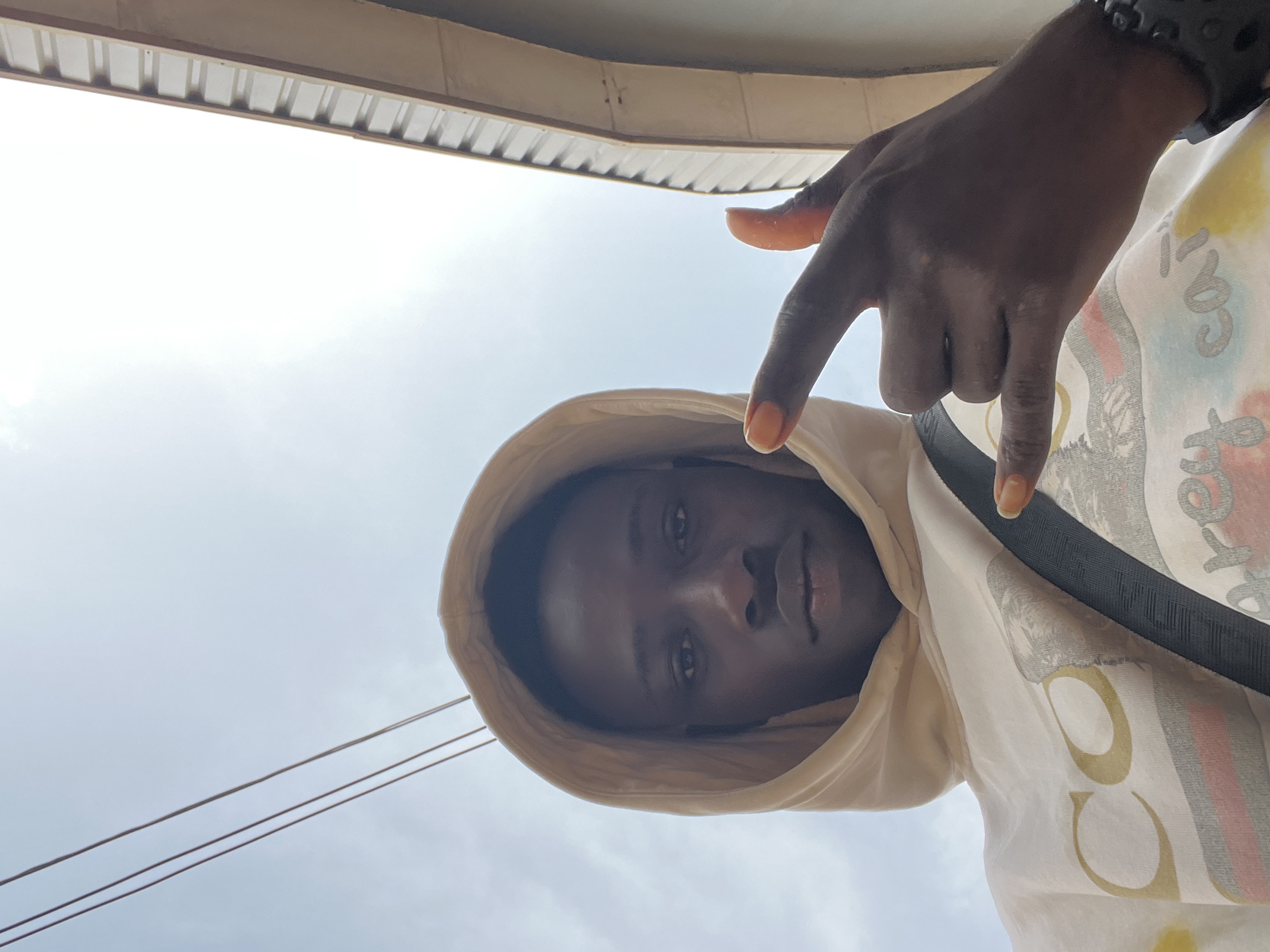 A low-angle, slightly distorted photograph taken outdoors in Obosi, Nigeria, captures a dark-skinned person wearing a hooded garment with a light tan hue. The subject's face, framed by the hood, is the focal point. The person's right hand, adorned with a black bracelet, is positioned above, fingers pointing downwards. The person wears a patterned white shirt with a black strap. Above the subject is a corrugated roof edge, and in the background, the sky is visible. Electrical wires cross the sky.