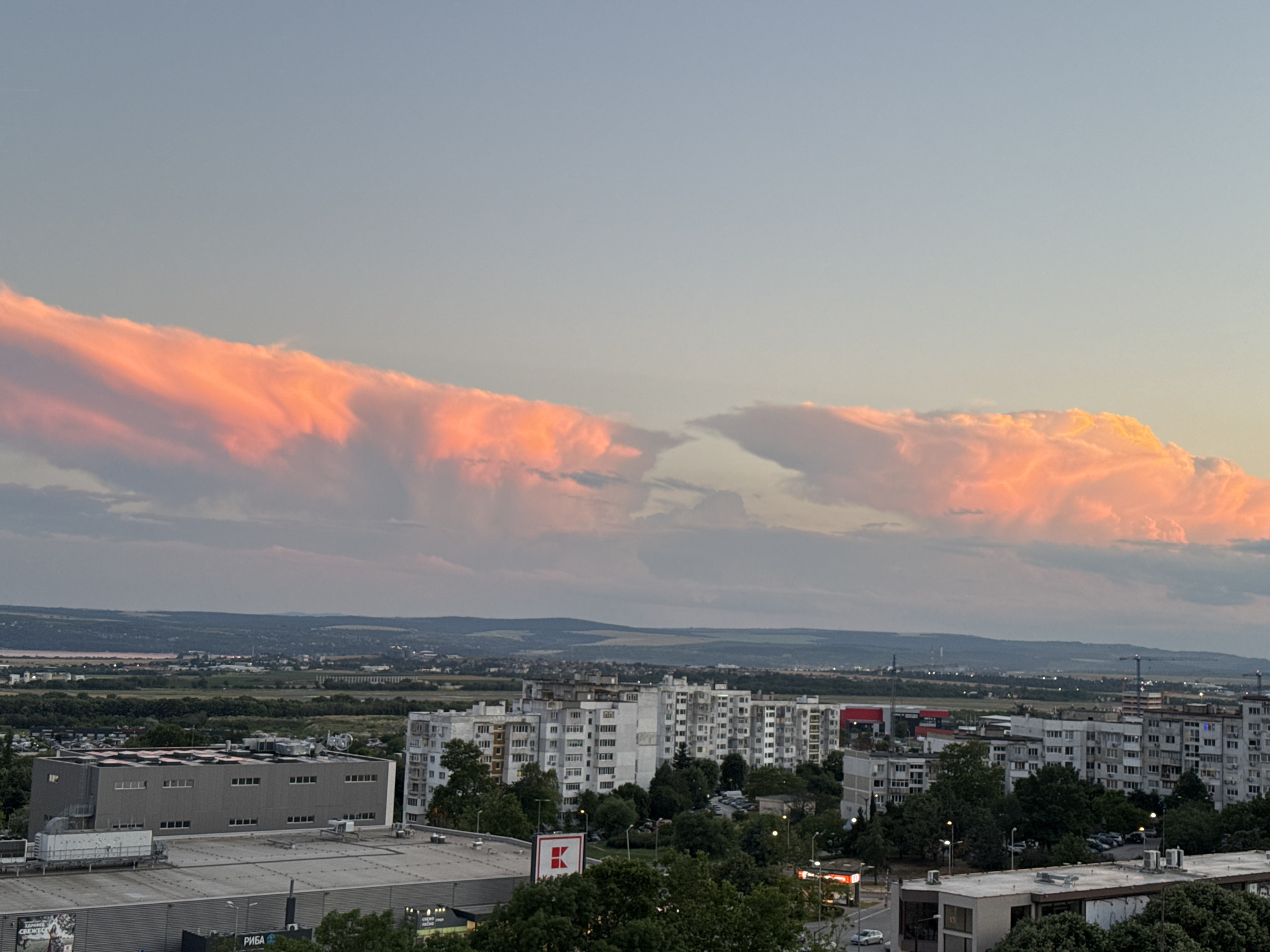 This is an aerial, landscape photograph of a city skyline at dusk, likely Varna, Bulgaria, featuring a dramatic sunset with vibrant orange and pink clouds. The foreground is dominated by a large, modern commercial building with a grey facade and a flat, white roof equipped with numerous HVAC units and ventilation systems. Adjacent to this, another building with a similar roof structure is visible, and part of a third building's rooftop with machinery is also present.
Further into the midground, a dense cluster of large, white, Brutalist-style apartment buildings stands out. These multi-story residential blocks feature numerous balconies and windows, characteristic of Soviet-era architecture. In front of these buildings, a prominent red and white logo, likely for a supermarket chain like Kaufland, is visible. Green trees partially obscure the lower levels of the apartment buildings and dot the urban landscape. Parking lots with several cars are scattered throughout the scene.
In the background, a vast, undulating landscape of rolling hills and agricultural fields stretches towards the horizon. A faint line of distant buildings and structures, possibly indicating further urban development or industrial areas, can be seen. The sky above is a pale blue, transitioning to a lighter hue near the horizon, with the striking sunset clouds occupying the upper portion of the frame. The overall impression is one of urban development set against a natural, scenic backdrop, illuminated by the dramatic colors of the setting sun.