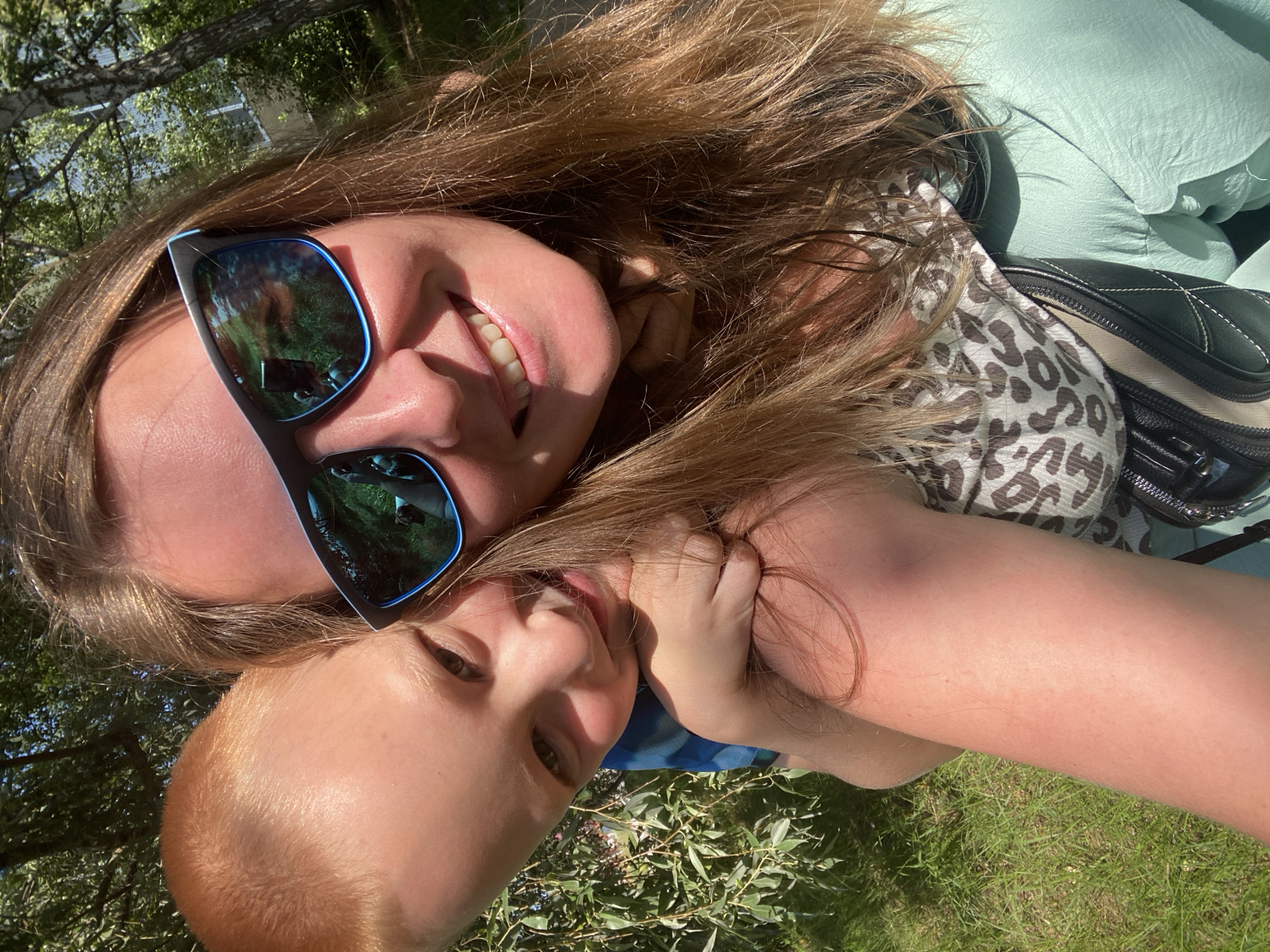 A close-up, selfie-style image captures a woman and a young child outdoors, with a backdrop of trees and grass. The woman, positioned on the right, is wearing sunglasses and smiling. Her long, brown hair is visible, and she appears to be holding the child. The child, on the left, has short, reddish-blonde hair and is looking at the camera, resting their head on the woman's shoulder. Both individuals are outdoors, in what appears to be a park or wooded area. The overall setting is sunny and appears to be a pleasant day. The image does not provide any specific geographic markers that would indicate its location within Noviy, Russia.