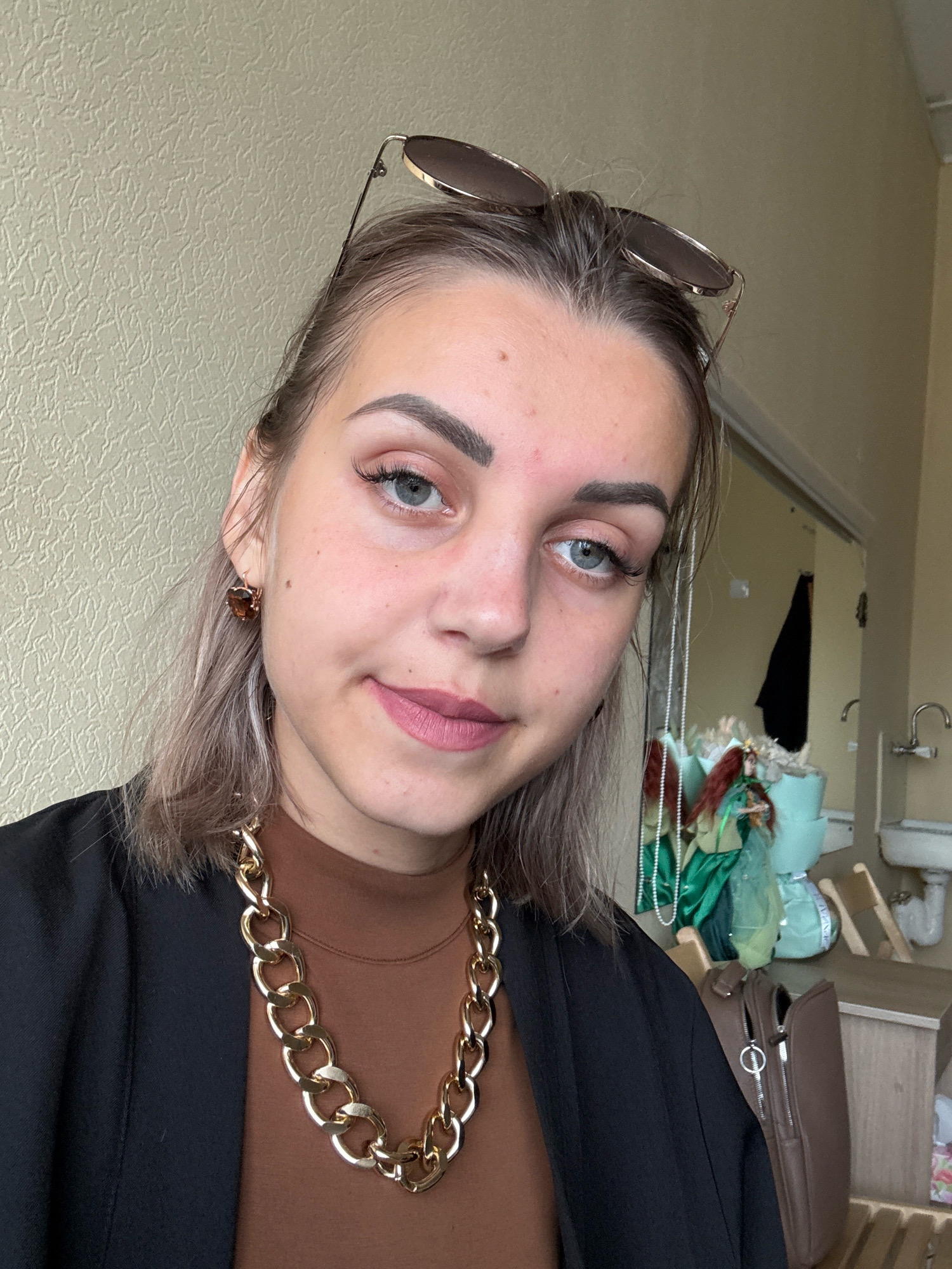 A young woman with light brown hair and light blue eyes is taking a selfie indoors. She is wearing a black blazer over a brown turtleneck and a thick gold chain necklace. Sunglasses are perched on top of her head. Her makeup is visible, including mascara and pink lipstick. In the background, a mirror reflects a closet with a black garment, a sink with faucets, and a bouquet of flowers. The wall behind her has a textured pattern. The overall setting suggests a domestic or possibly a studio environment, but there are no explicit indicators of the city of Yuzhno-Sakhalinsk, Russia.