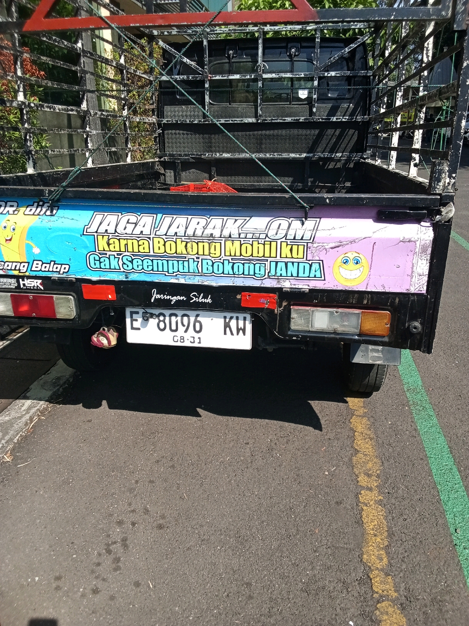 The image shows the rear of a white pickup truck parked on an asphalt road with yellow and green painted lines. The truck has a black metal cage over its bed. The rear of the truck features several stickers with Indonesian text and cartoon graphics. One sticker, with a blue background, reads "JAGA JARAK .OM Karna Bokong Mobil Ku Gak Seempuk Bokong JANDA". Another sticker features a cartoon banana character and the text "Sang Balap". A third sticker has a smiling emoji with blue eyes. The truck has a license plate that reads "E 8096 KW 08-31". A single sandal is hanging from the bumper. The truck appears to be in a parked position on a street. The background shows foliage and a building. The lighting suggests it is daytime.