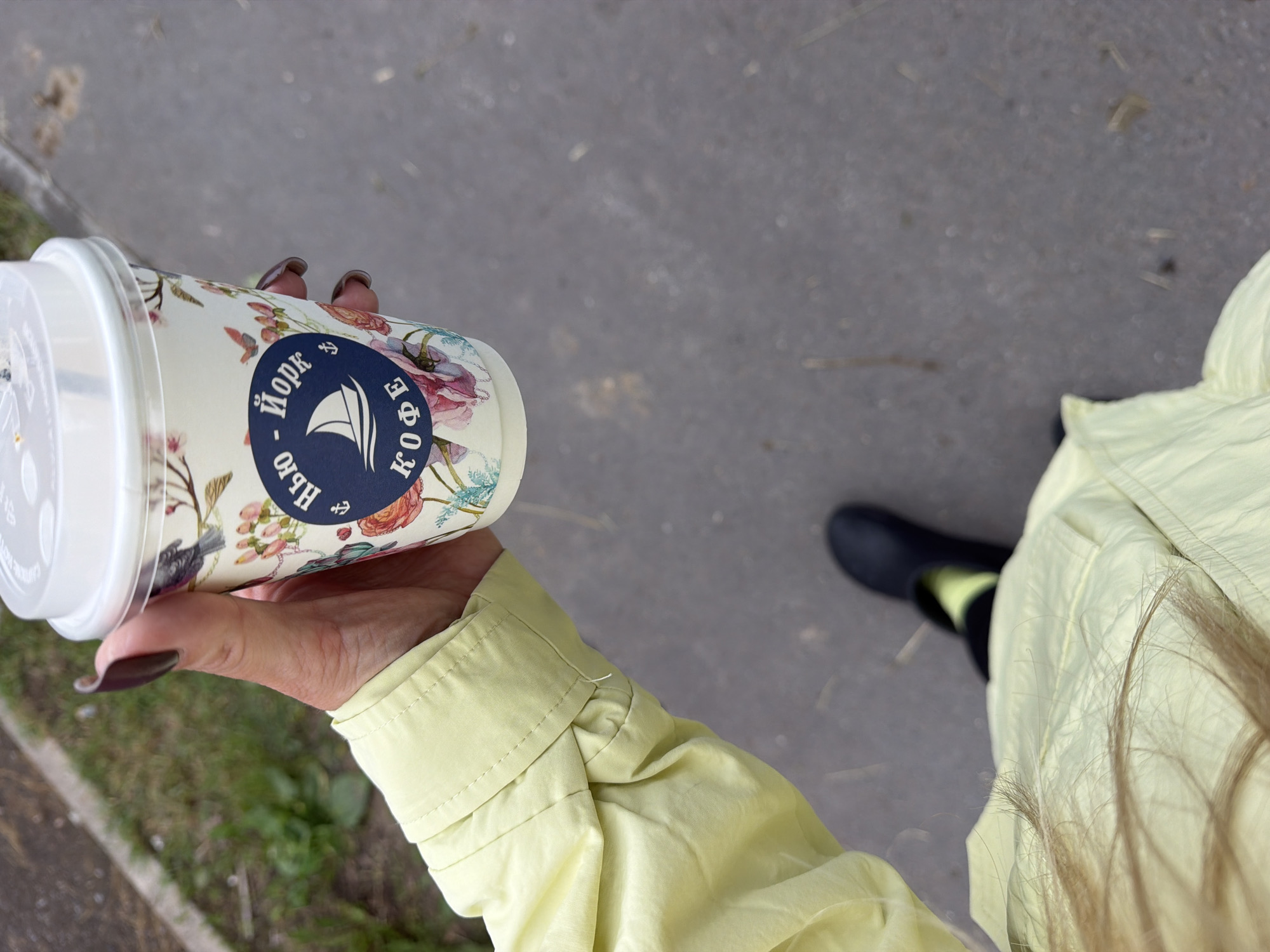 A close-up, overhead shot captures a person's hand holding a floral-patterned disposable coffee cup. The cup features a dark blue circular logo with a sailboat and the text "Нью-Йорк КОФЕ" in white lettering. The person is wearing a light yellow jacket with prominent stitching on the cuff. A dark rubber boot is visible on the ground below, paired with a bright yellow sock. The setting appears to be an urban environment, with a paved surface and a grassy area in the background. The presence of a coffee cup and urban pavement suggests a location such as Votkinsk, Russia.