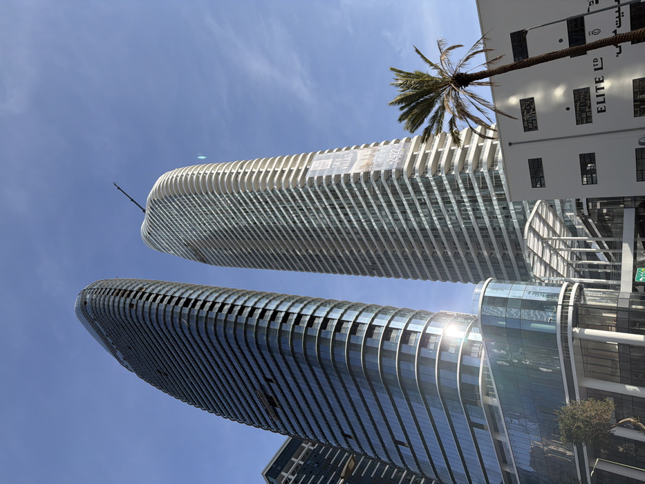The image depicts an urban architectural scene in Tel-Aviv, Israel, specifically showcasing the Midtown complex. Two prominent, modern, curved skyscrapers dominate the view, observed from a low angle against a clear blue sky.
The skyscraper on the left features a dark blue-tinted glass facade with visible horizontal bands and some incomplete sections suggesting ongoing construction or finishing work, particularly on its lower levels where sunlight reflects intensely. Its base incorporates a multi-story glass-clad podium.
The skyscraper on the right presents a lighter facade composed of numerous horizontal white or light-colored balcony bands interspersed with large glass windows, also with a distinct curved profile. A large vertical banner affixed to its side advertises "SKY VILLAS" and a numerical string "-2422". A construction crane arm is visible at its apex, extending against the sky.
In the mid-ground, between the two towers, a contemporary elevated walkway or connecting structure with white, open beamwork is visible. To the far right, a tall palm tree with a slender trunk and green fronds leans into the frame. Beneath the palm tree and to the right, a white multi-story building with rectangular windows is partially visible. This building features a black sign bearing the text "ELITE LTD" in English, with additional Hebrew text below it.
The sky is uniformly clear and blue, indicating bright daylight conditions. No individuals or discernible actions are present within the frame. The specific location is the Midtown Tel Aviv complex, visible from street level near Menachem Begin Road.