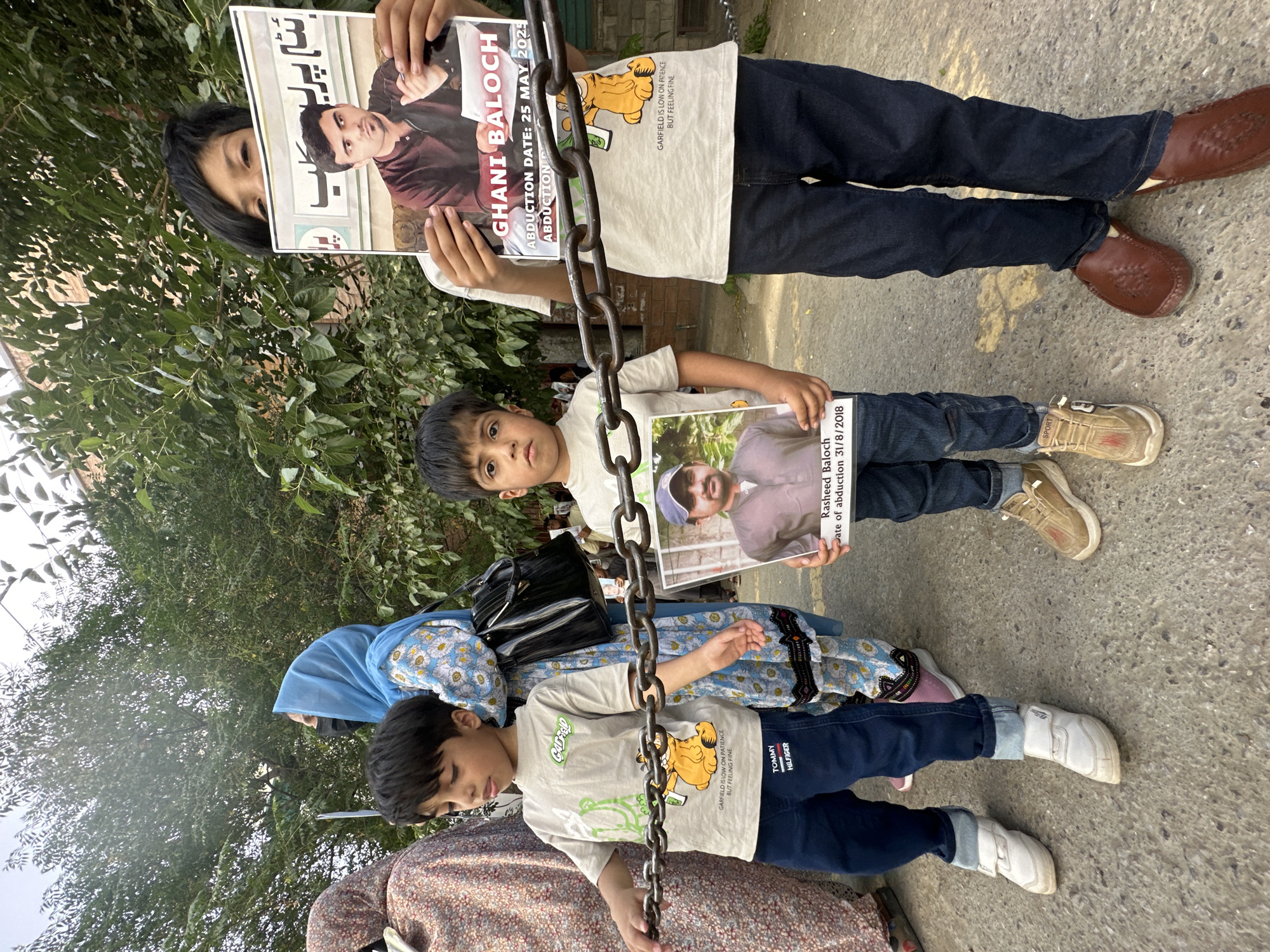 Three children are standing on a paved surface outdoors, likely a sidewalk or street. They are holding printed posters displaying photos and names of individuals identified as "Ghani Baloch" and "Rasheed Baloch" along with their respective abduction dates. A metal chain hangs in front of the children. An adult in a floral dress and blue hijab is partially visible at the edge of the image. The environment suggests a residential or public area with trees and foliage in the background. The text on the posters is in English and Urdu. One of the children is wearing a Tommy Hilfiger labeled pants. The Garfield cartoon character is printed on two of the children's t-shirts. The location is presumed to be Quetta City Tehsil, Pakistan.