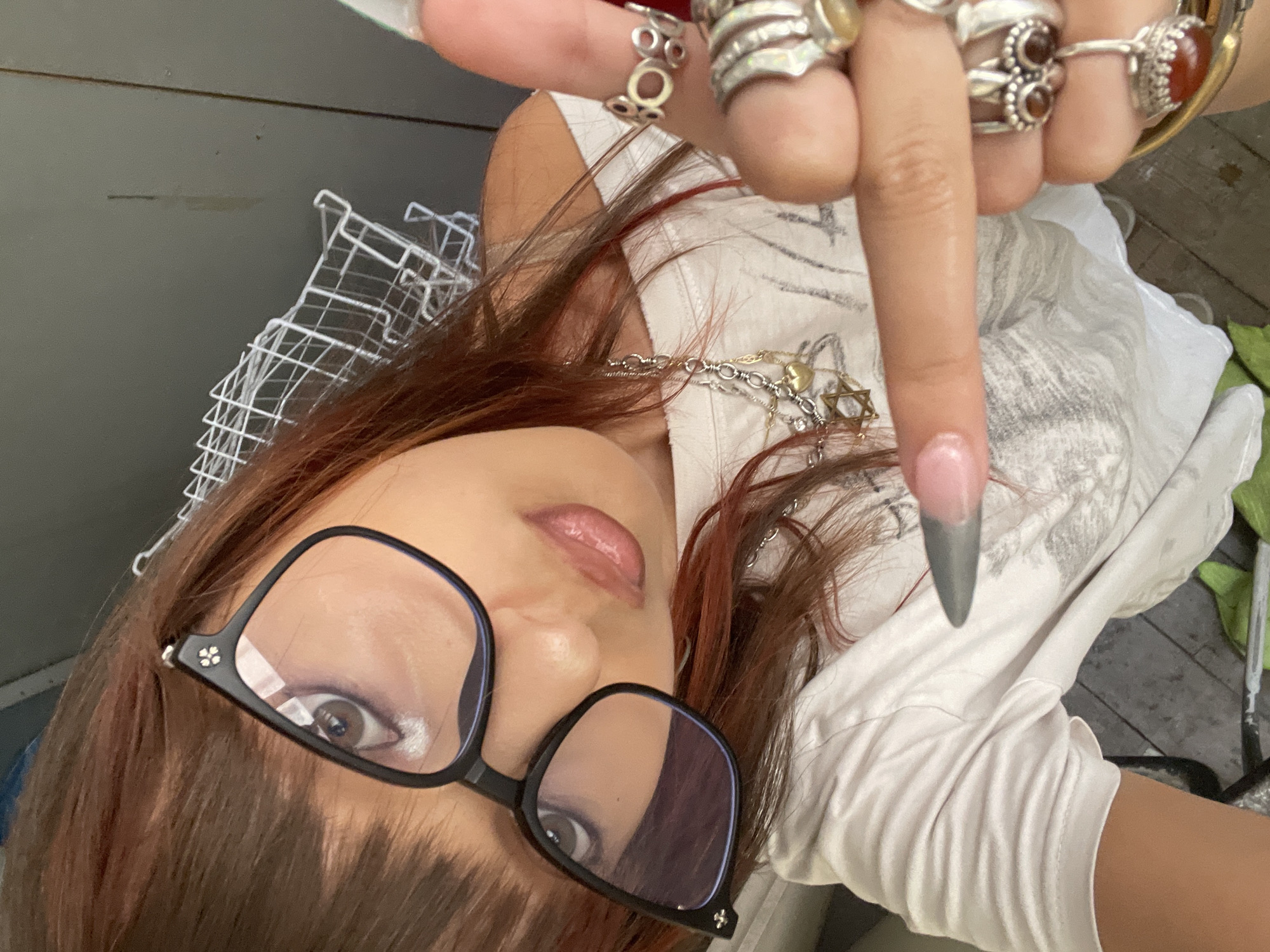 The image shows a close-up of a young person wearing black-rimmed glasses and multiple rings on their fingers. The person has reddish-brown hair with bangs and is wearing a white t-shirt with a graphic print. A silver necklace with a star pendant is visible. The person is pointing their right index finger towards the camera, and their left hand is also adorned with rings. The background appears to be an indoor or semi-outdoor environment with a grey wall and some wire shelving. There is no discernible information within the image that places the scene in Raanana, Israel.