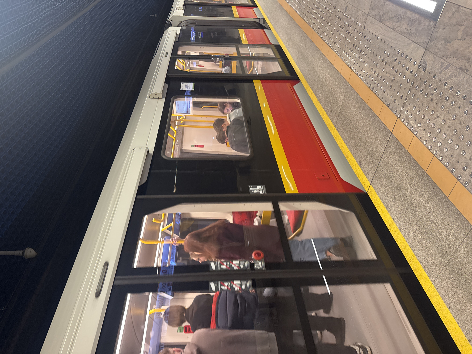 An underground metro station scene shows a train stopped at a platform, viewed from an angled perspective looking along the side of the train. The train, positioned on the left side of the frame and running vertically, features a distinctive livery of red along its lower body, a prominent yellow stripe above it, and black window sections with white trim. Multiple cars of the train are visible, extending into the distance.
Inside the train, several passengers are visible through the windows. They appear to be standing or seated, with some individuals looking at their mobile phones. Reflections on the glass also show parts of the interior and distorted views of the platform.
To the right of the train is the station platform, constructed with light grey, speckled tiles. A clear yellow safety line runs along the platform's edge, adjacent to the train. Further back on the platform, tactile paving with circular raised dots is present, common in public transport areas.
On the far left of the image, serving as the background opposite the platform, is the dark wall of the metro tunnel. It features a repeating geometric pattern, possibly a grid of squares, adding texture to the otherwise dark space.
The scene is illuminated by artificial lighting, consistent with an underground setting. The train's red and yellow coloring is characteristic of the Warsaw Metro M1 line in Poland. A small, white rectangular sign is affixed to one of the train windows, but its text is illegible due to distance and blur. No specific activity beyond the train being stationary at the platform is clearly depicted.