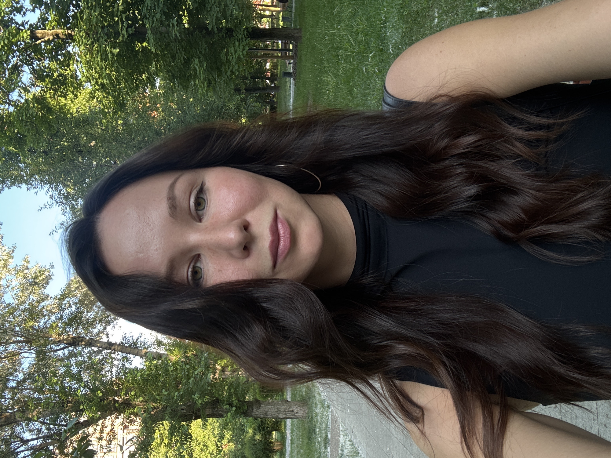 The image is a selfie of a young woman, appearing to be taken in a park in Moscow, Russia. She has long, wavy dark brown hair that cascades around her face and over her shoulders. Her eyes are light brown or hazel, and she looks directly at the camera with a calm, slightly smiling expression. She wears natural-looking makeup, including light pink lipstick, and a black top. A subtle hoop earring is visible on her right ear.
The background is a vibrant green park setting. Numerous trees with lush foliage are visible, indicating good weather and a sunny day, with sunlight dappling through the leaves. Behind her, there's an expanse of green grass and a paved pathway or road, further suggesting a public park or green space. No visible text is present in the image.