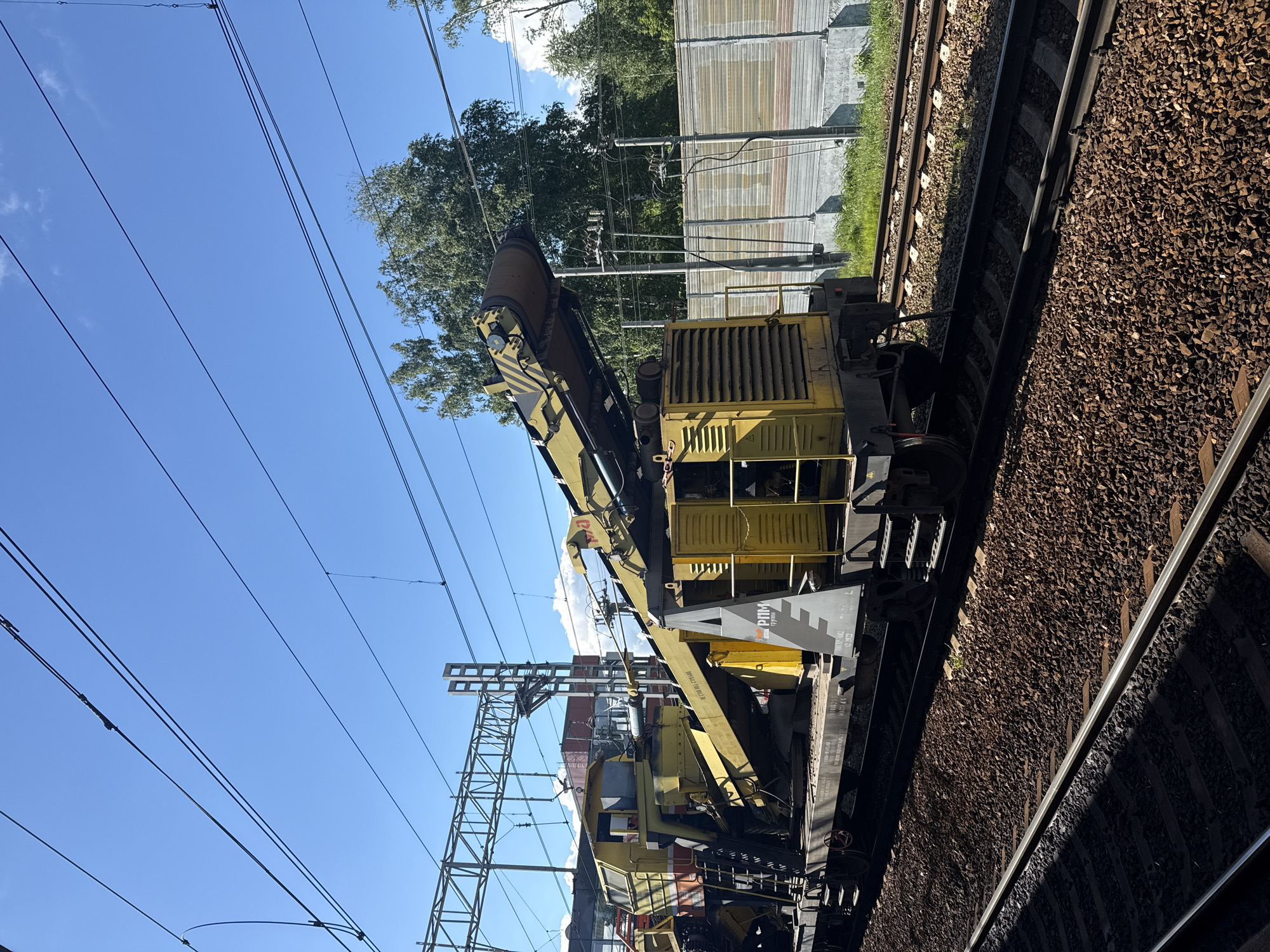 The image shows a large, yellow rail maintenance vehicle positioned on a train track. The surrounding environment includes track ballast, overhead power lines, a fence, and foliage suggesting an outdoor setting. The location is not discernible from the image.