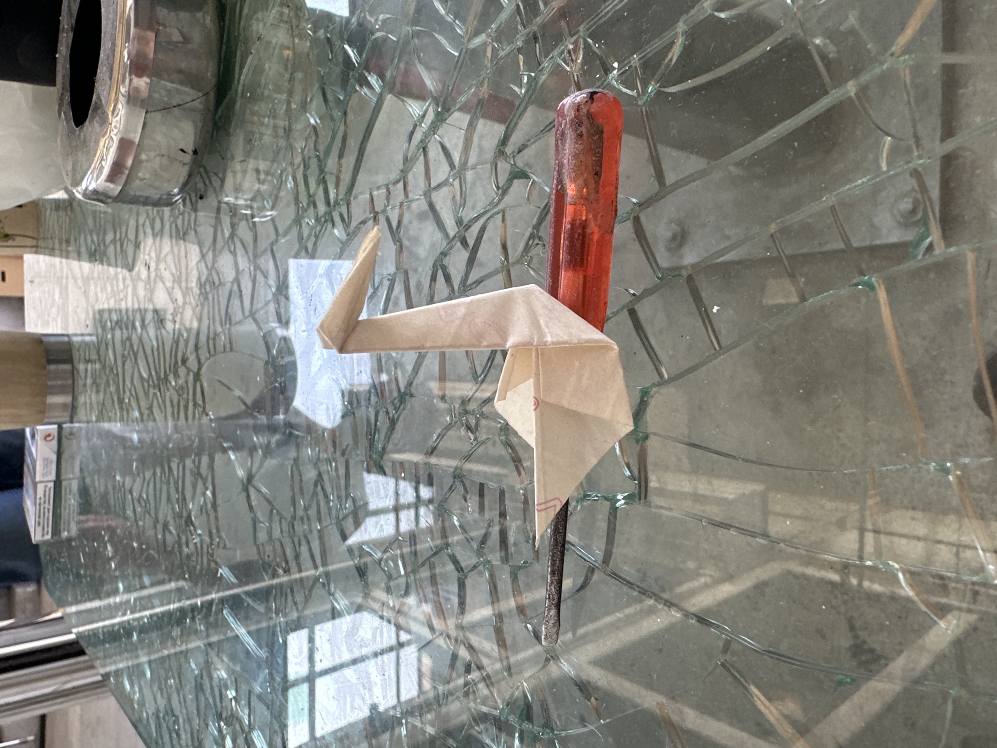 A light brown origami crane is placed on a broken glass table. The crane is positioned horizontally, with its beak pointing to the left. Underneath the crane's body is a red object, possibly a tool handle or a pen. The table's glass surface is shattered into many pieces, creating a web-like pattern. In the background, a metallic ashtray with a circular opening is visible. The lighting suggests an indoor environment, possibly outdoors given the shadows. The exact location is unknown.