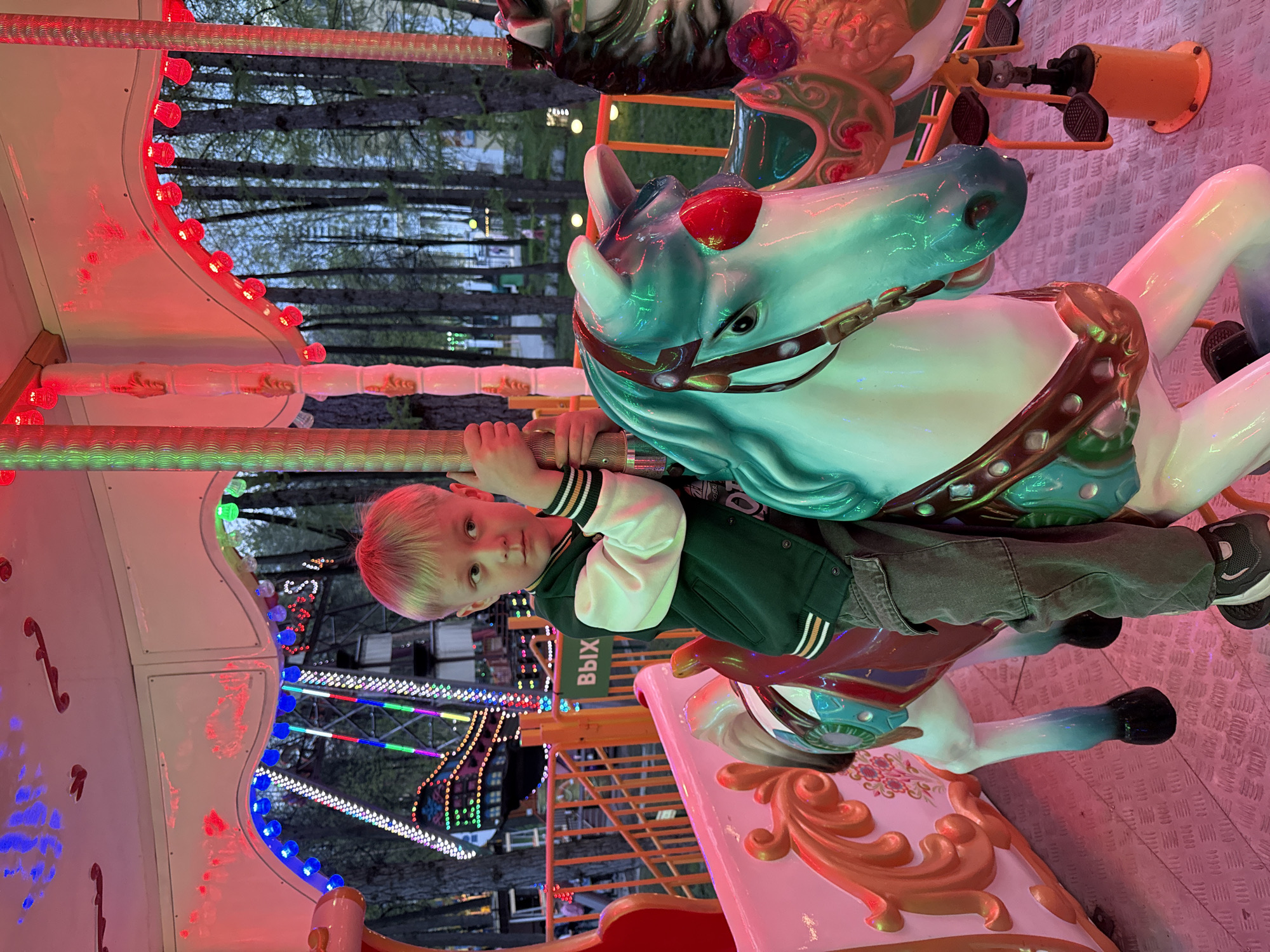 A boy with blond hair is riding a carousel horse. The carousel is brightly lit with red, green, and blue lights. In the background, a Ferris wheel is visible. A sign with Cyrillic letters "ВЫХ" is placed vertically on the carousel. The image was taken in Perm, Russia.