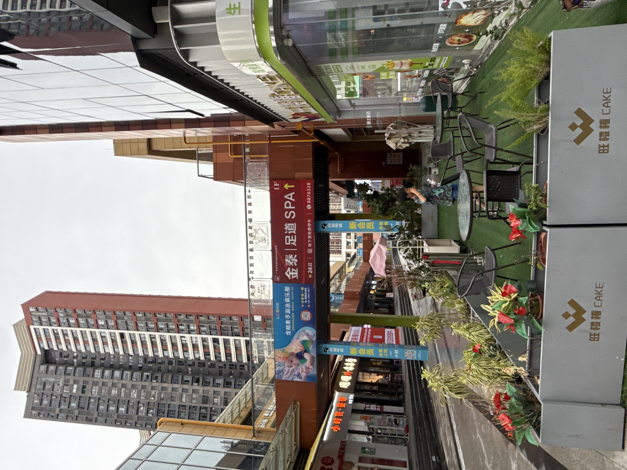 The image captures a street-level view of a commercial area in Huizhou, China, with a focus on the outdoor seating area of a cafe or restaurant. In the foreground, there are grey planters containing artificial red flowers and green foliage, with a logo featuring a stylized 'W' and Chinese characters that translate to "CAKE" on the front planter. A few black metal chairs and round tables are arranged on an artificial green lawn, suggesting an outdoor dining or seating space.
Behind the seating area, a commercial building with large glass windows and signage is visible. One sign features green lettering and the word "CAKE" along with other Chinese characters. Further back, a prominent red and grey high-rise apartment building dominates the skyline, characteristic of urban development. In the middle ground, a pedestrian walkway or street is visible, with other shops and businesses lining the sides. An overhead walkway with advertisements connects buildings, and one large banner promotes a swimming club and spa. A young boy wearing a patterned shirt is standing and looking towards the right side of the image, holding what appears to be a camera or phone. The sky is overcast, indicating a cloudy day. The overall scene depicts a modern urban commercial district with a mix of residential and retail spaces.
