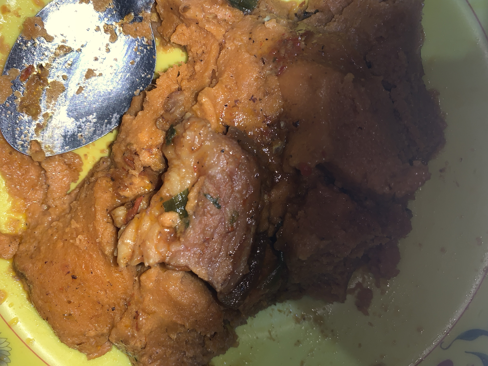 A plate of food, possibly yam pottage, is on a yellow plate with a silver spoon. The food appears to be a mixture of mashed yam and stew, containing pieces of meat or fish, as well as seasonings, in an indistinct indoor environment. The location is New Stadium, Nigeria.