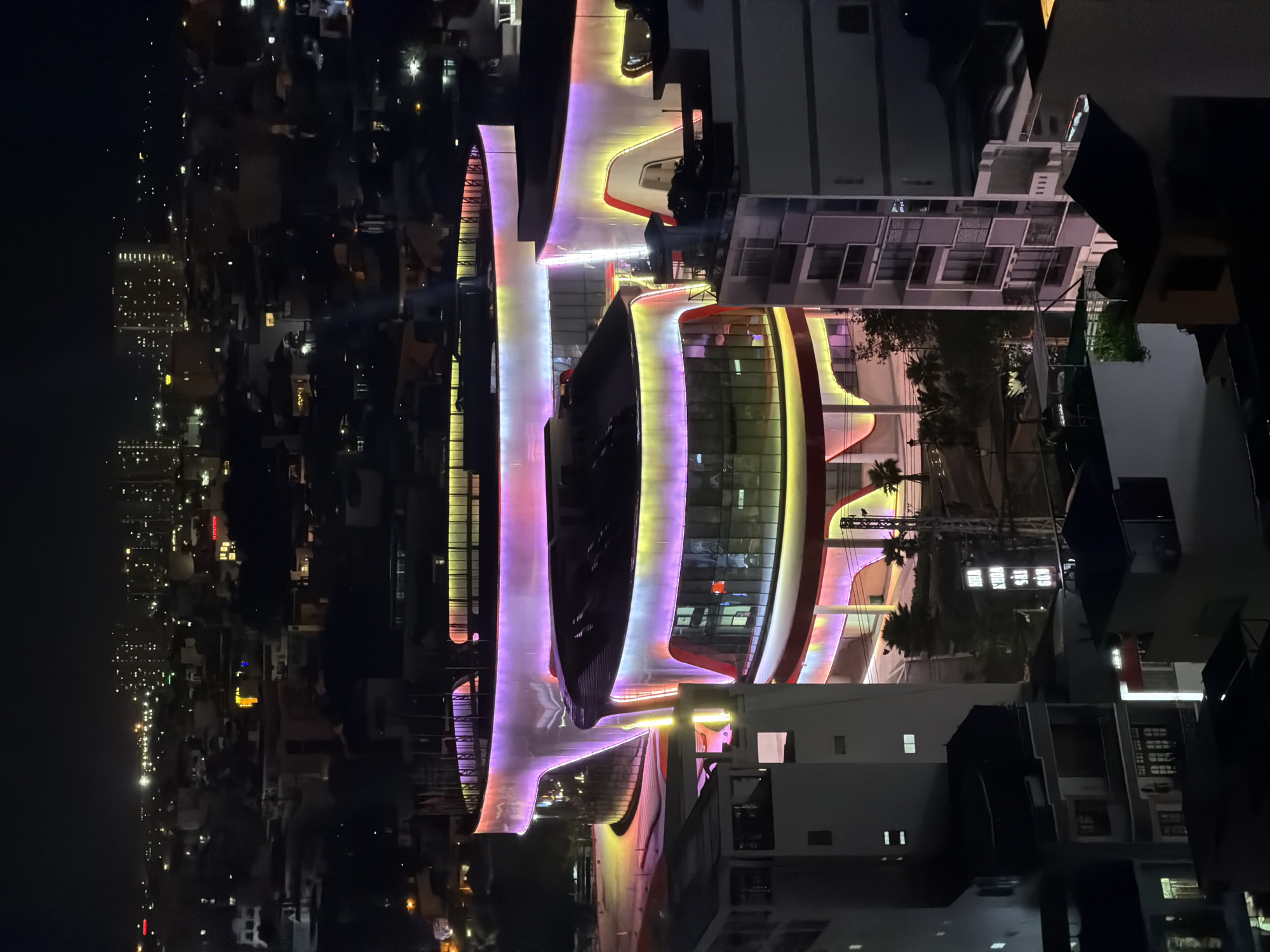 This is a night aerial shot of a modern, brightly lit building in Nha Trang Ward, Vietnam. The building features a distinctive curved, multi-level design with smooth, white exterior walls illuminated by vibrant, neon-like lights in shades of purple, pink, and yellow. These lights trace the contours of the building, creating a dynamic and futuristic aesthetic. The building is surrounded by other, less illuminated structures, some appearing to be residential or commercial buildings, and is set against a backdrop of a city skyline with numerous distant lights. Palm trees are visible in the foreground, suggesting a tropical or subtropical location. The overall impression is one of a modern architectural landmark in an urban environment at night.