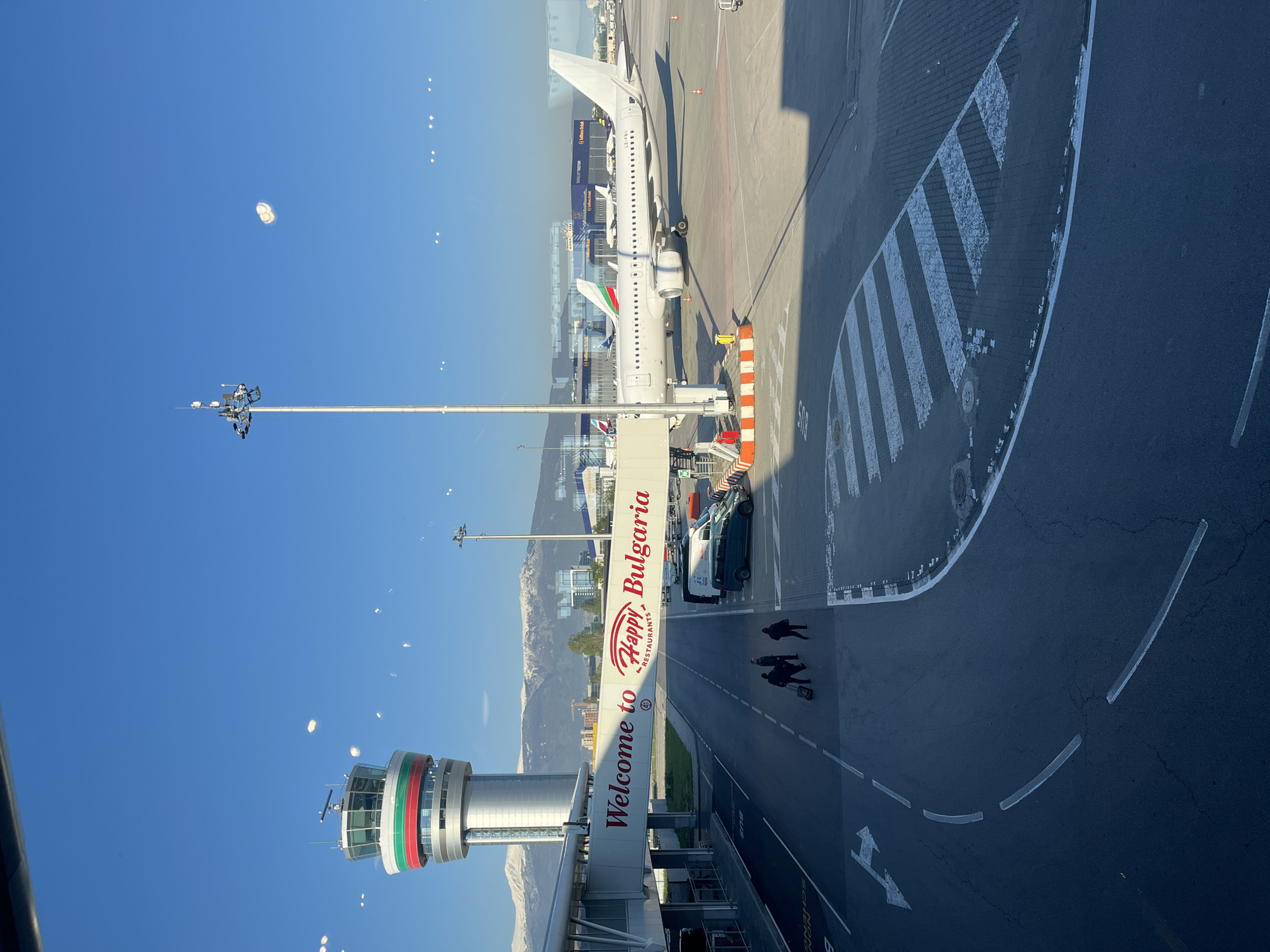 The image depicts a view of Sofia Airport in Bulgaria, seen from an elevated perspective, possibly from within the airport terminal.
In the foreground, there is an airport control tower with horizontal stripes of red, white, and green – the colors of the Bulgarian flag. A pedestrian walkway with the words "Welcome to Bulgaria" and the "Happy Restaurants" logo bridges the tower to another building.
An airplane, likely an Airbus A320 family aircraft, sits at the gate with a boarding bridge attached. Ground vehicles, including buses, are visible alongside the aircraft. People, possibly passengers, are walking on the paved area towards the terminal.
The airport apron is marked with directional signage and striped zones. In the background, there are buildings, likely airport facilities, and distant mountain ranges.
The sky is clear and blue.