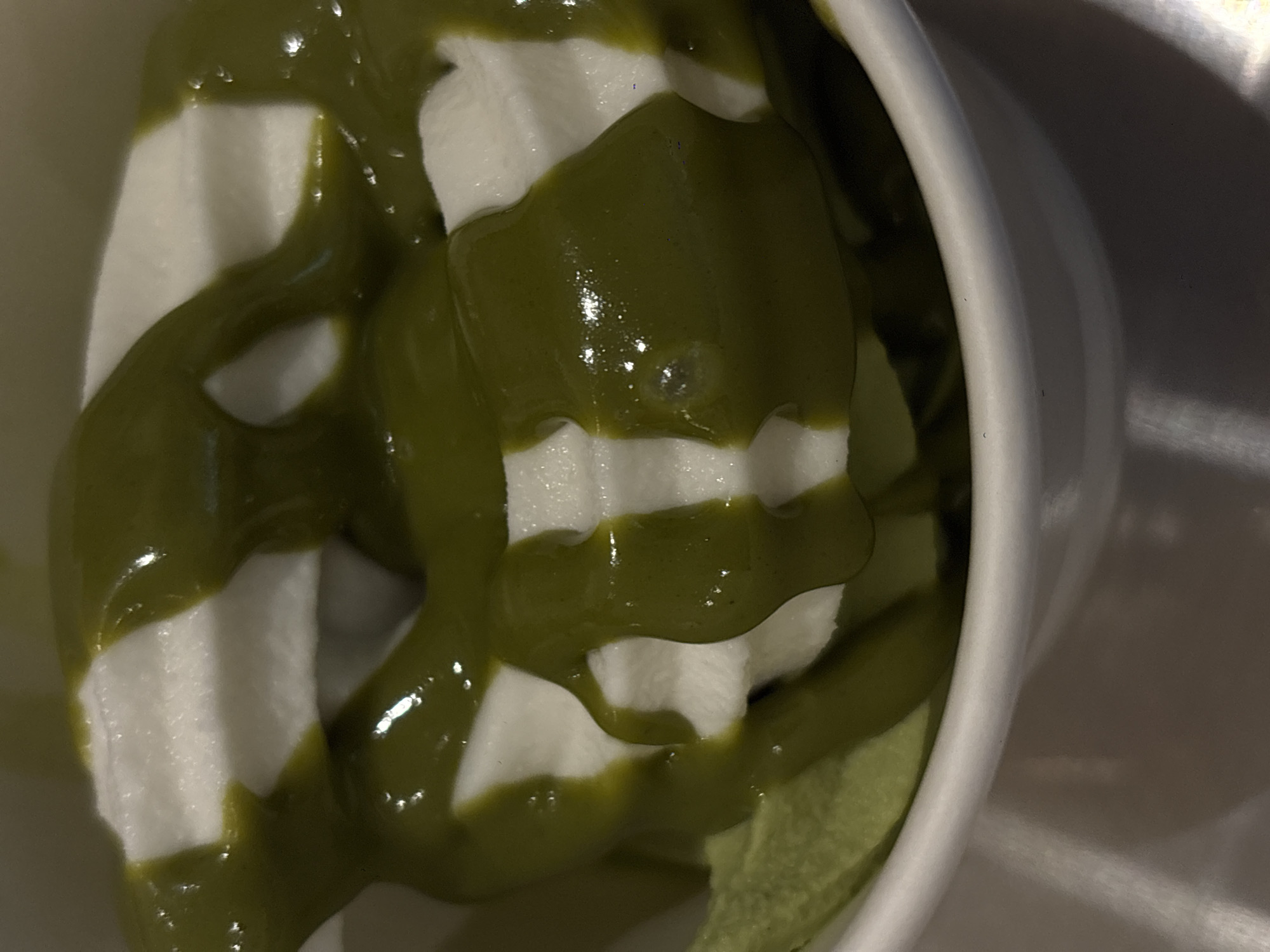 A high-angle, close-up shot depicts a white ceramic cup filled with soft-serve ice cream drizzled with green matcha sauce. The ice cream appears swirled, its white color contrasting with the dark green of the matcha. The cup is situated on a light beige or off-white surface. The scene is brightly lit, highlighting the textures and colors of the dessert. There is no evidence to suggest this photo was taken in Mexico City, Mexico. No people, actions, interactions or further environmental details are present.