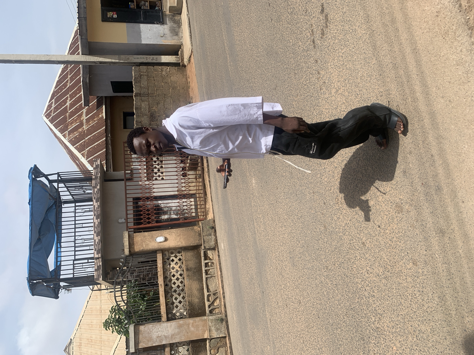 A young man stands on a paved road in Barakin Akawo, Nigeria. He is wearing a white jacket and black pants, and sandals. He is holding a selfie stick in his right hand. Behind him are buildings of varying construction styles, some with tin roofs and wrought iron balconies. The road is asphalt. The lighting suggests it is daytime.