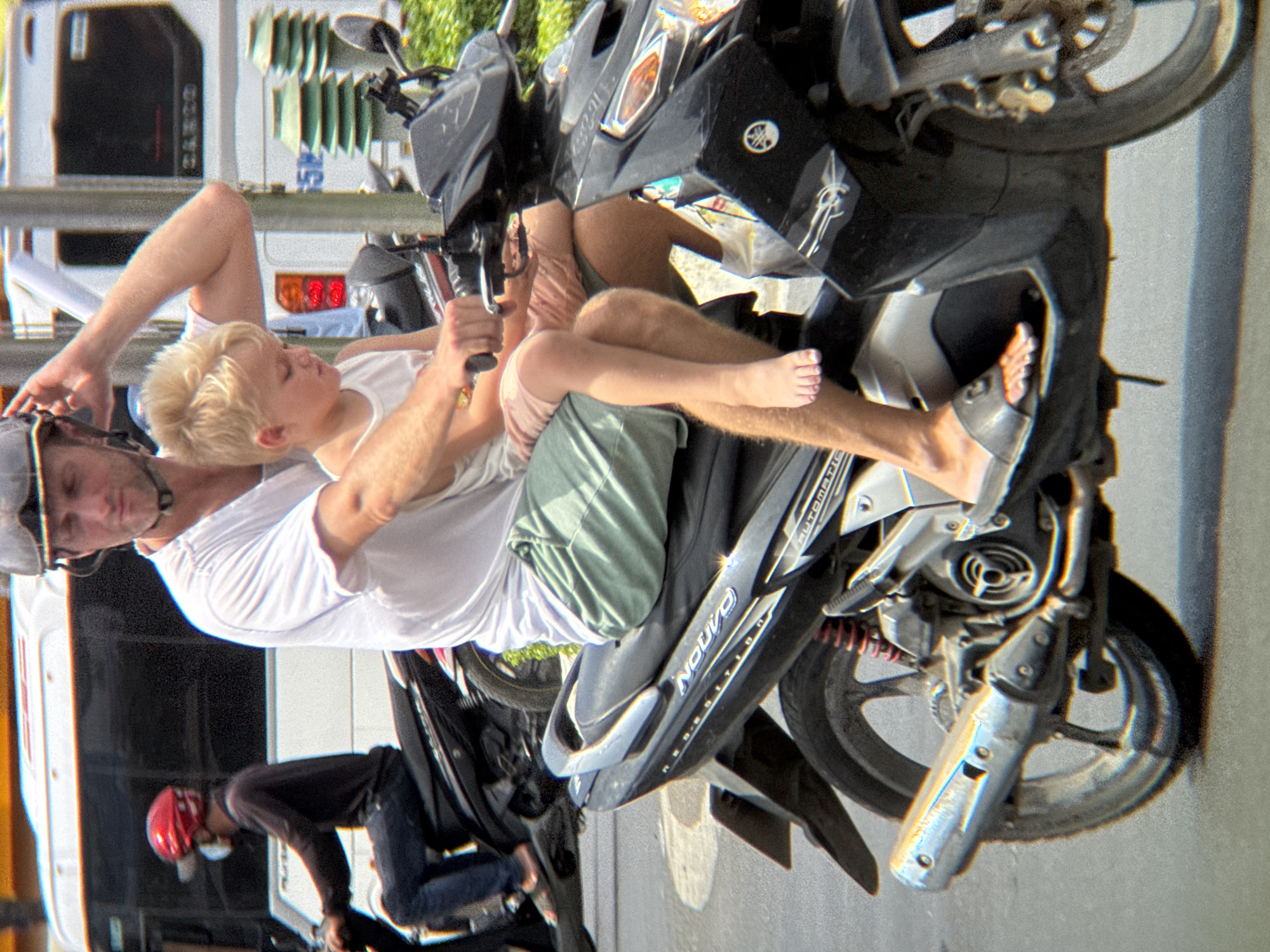 This image depicts a sunny outdoor scene in Nha Trang, Vietnam, focusing on a man and a child riding a black scooter. The man, with fair skin and a shaved head, wears a grey helmet and a white t-shirt. He is holding the handlebars of the scooter with his right hand. A young boy with blond hair, wearing a white tank top and pink shorts, is seated in front of the man. The boy's left leg is visible, dangling in front of the man's leg. Both are wearing casual attire.
The scooter is a dark color, with visible chrome accents and a silver exhaust pipe. The "NOVO" branding is prominent on the side of the scooter. In the background, a white bus with tinted windows and a red and white striped awning is partially visible. Another scooter with a rider wearing a red helmet and a dark top is visible further back. The environment suggests a street scene with some greenery visible on the right side of the frame, and what appears to be a sidewalk or road surface in the foreground. The overall lighting indicates it is daytime with bright sunlight.