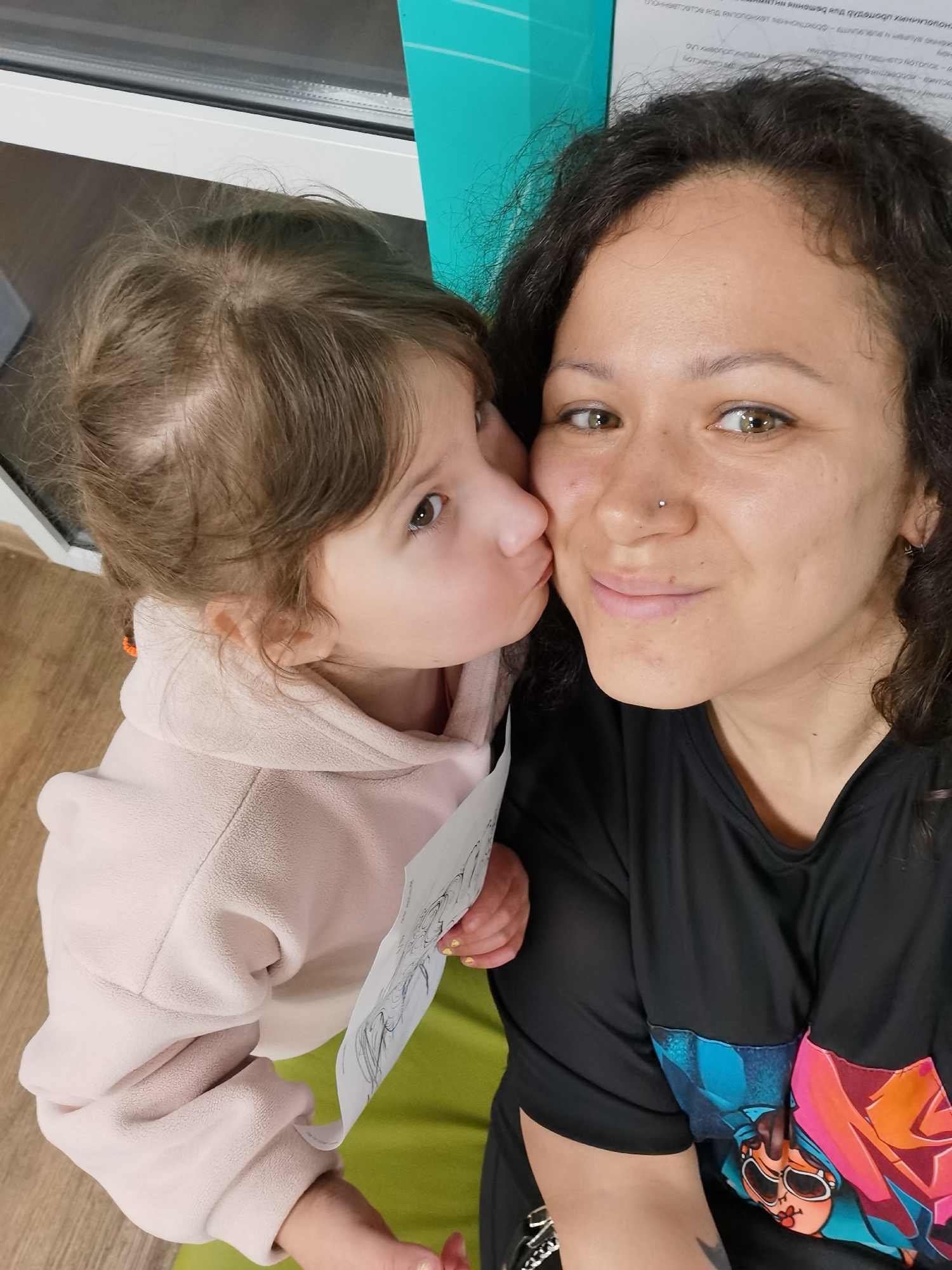 The image captures an adult female and a young child in an indoor setting. The adult female, appearing to be in her 20s or 30s, has dark, curly hair, olive skin, and greenish-brown eyes. She wears a small silver stud nose piercing on her left nostril and a small silver hoop earring in her left ear. Her fingernails are painted yellow. She is smiling softly towards the camera and wearing a black short-sleeved t-shirt with a vibrant, colorful graphic print depicting an animated character on the lower front. A partial tattoo is visible on her inner right arm.
The child, a toddler or very young child likely between 2 and 4 years old, has light brown hair with visible patches of thinner hair on the scalp. She has brown eyes and is seen leaning in from the left to kiss the adult female's cheek and nose area with pursed lips. She is wearing a light pink hooded fleece pullover. A small orange accessory is visible near her left ear. Both the child and the adult appear to be holding a white piece of paper with a black and white drawing on it.
The background features a white-framed window on the left. To the right of the window, there is a vertical panel of light teal or aqua color. Above this panel, a white sign or document is visible with text in Cyrillic script. The legible text includes phrases such as "ФЕДЕРАЛЬНОЕ ГОСУДАРСТВЕННОЕ БЮДЖЕТНОЕ ОБРАЗОВАТЕЛЬНОЕ УЧРЕЖДЕНИЕ ВЫСШЕГО ОБРАЗОВАНИЯ" (Federal State Budgetary Educational Institution of Higher Education) and "МИНИСТЕРСТВО ОБРАЗОВАНИЯ И НАУКИ" (Ministry of Education and Science), indicating a Russian educational institution. The specific city of Izhevsk is not identifiable from the visible details within the image. The floor on the left is light brown wood, and a bright green surface, possibly a cushion or mat, is situated below the child.