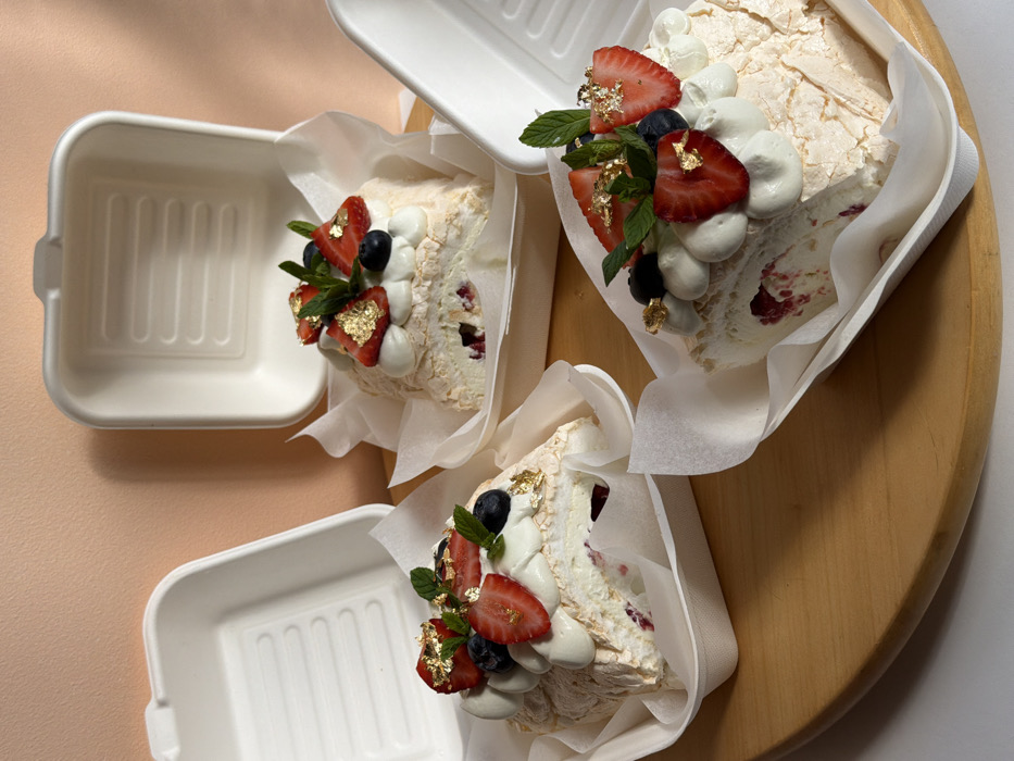 The image displays three individual dessert portions, likely meringue rolls or pavlovas, prepared for take-away in off-white, compostable molded fiber containers. Each dessert consists of a textured, light-colored meringue shell encompassing a creamy white filling with visible dark red fruit inclusions. Garnish on each includes sliced fresh strawberries, whole blueberries, sprigs of green mint, and scattered gold leaf flakes. Two containers have their matching hinged lids open, positioned upright behind the desserts. All items are arranged on a light brown, smooth wooden surface, set against a plain, light peach-toned wall. The scene is located in Tel-Aviv, Israel.