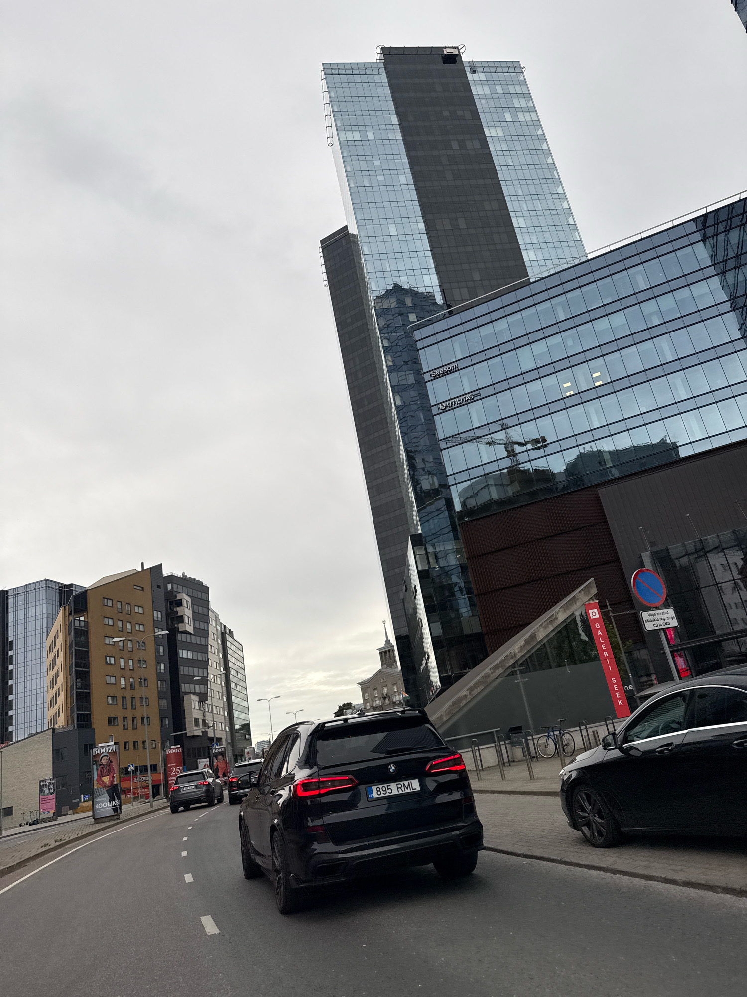 In Tallinn, Estonia, on an overcast day, a street scene unfolds with modern architecture and traffic. A black BMW X5, license plate 895 RML, is in the foreground, its rear lights illuminated, driving on a grey asphalt road. To its right, a black Mercedes-Benz sedan is parked on the sidewalk. The road is marked with white dashed lines. In the background, a series of contemporary buildings dominate the skyline. On the left, a cluster of multi-story buildings includes a yellow and brown structure with numerous windows. To the right, a very tall, angular skyscraper with a glass facade and dark vertical accents rises dramatically. A lower, wider glass-fronted building adjoins it. The sky is a uniform grey. Signs visible on the buildings and street include "Booz," "Kooliks," "Galerii Seek," and a blue circular sign with a red bar indicating a prohibition. A bicycle is parked near the buildings. A construction crane is reflected in the glass facade of one of the skyscrapers.
