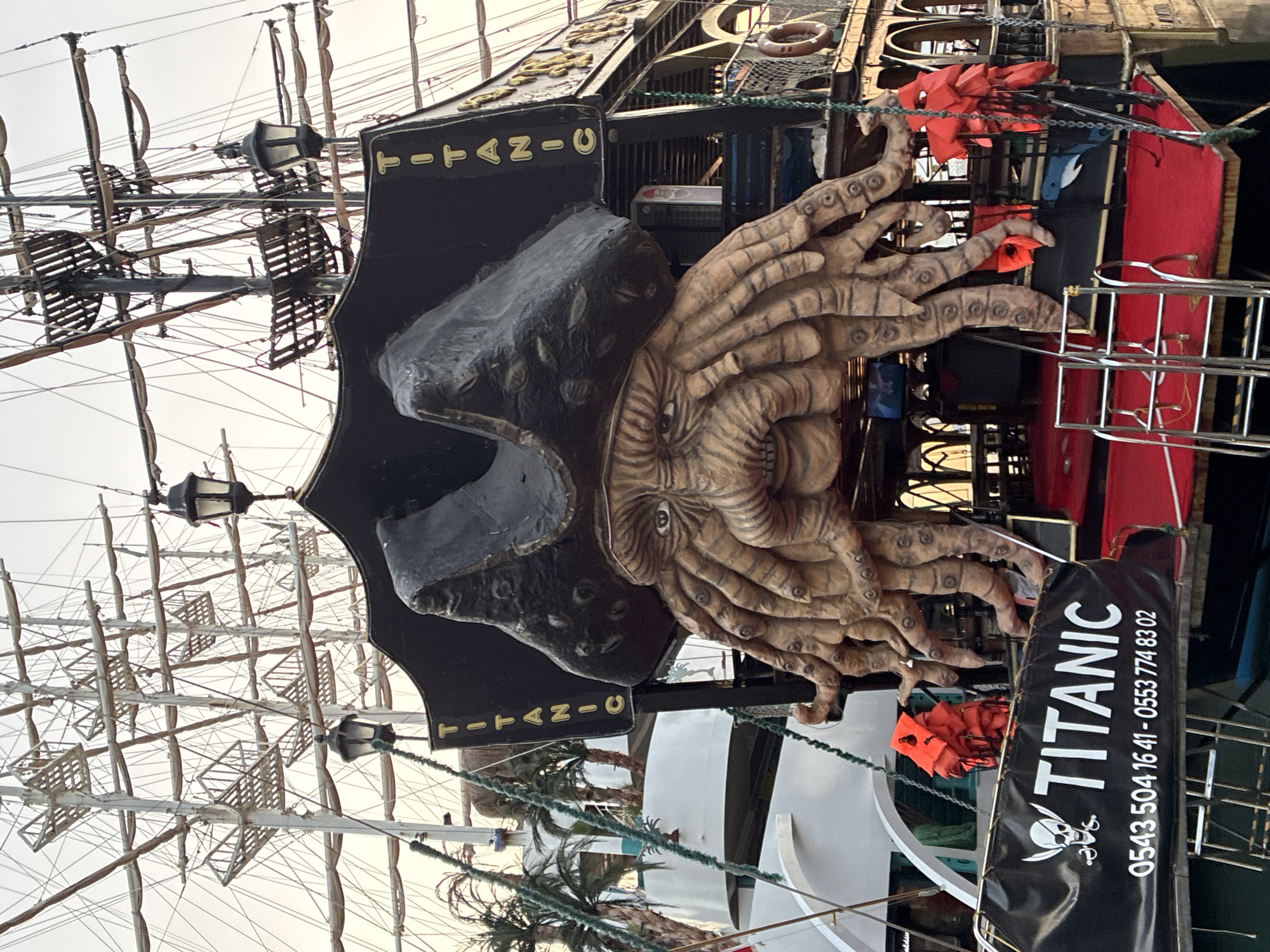 The image shows the stern of a pirate-themed boat named "Titanic" docked in Alanya, Turkey. The boat features a large, grotesque sea creature figure with a pirate hat on its head, serving as the main decoration. Several orange life vests are visible, some attached to the boat's railing and others stacked. The boat's name "Titanic" and a phone number are prominently displayed on a black banner at the back. In the background, the masts and rigging of other boats and the sky can be seen. The overall scene depicts a recreational boat ready for passengers.
