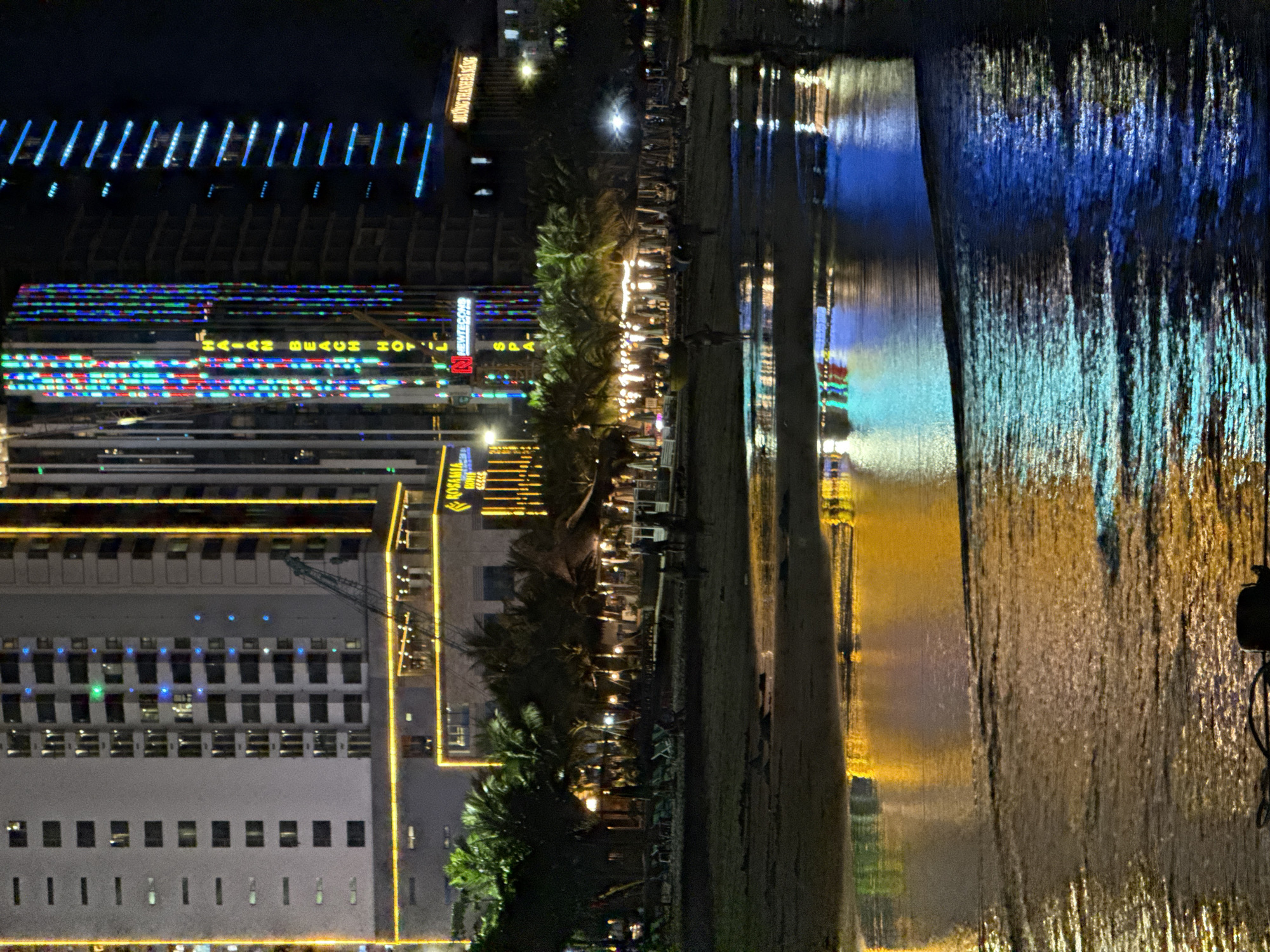 The image captures a nighttime scene on a beach in Da Nang, Vietnam, characterized by the vibrant reflections of illuminated buildings on the wet sand and gentle waves. In the background, towering modern buildings are adorned with colorful LED lights, creating a striking visual contrast against the dark sky. One building displays vertical stripes of red, blue, and green lights, with the words "HAIAIN BEACH HOTEL" and "NEWTECONS" visible. Another building features a series of horizontal blue lights. To the left, a building with a grid of windows is outlined with yellow lights, and a crane is positioned in front of it.
In the mid-ground, a strip of palm trees separates the buildings from the beach. Beach umbrellas and lounge chairs are set up, indicating a resort area. Several indistinct figures, appearing as silhouettes, are scattered across the beach, some walking and others appearing to be stationary.
The foreground is dominated by the wet sand and shallow water, where the lights from the buildings are reflected in dazzling patterns of yellow, blue, and green. The reflections are distorted by the movement of the water, creating abstract streaks of color. The overall atmosphere is one of a lively, modern coastal city at night.