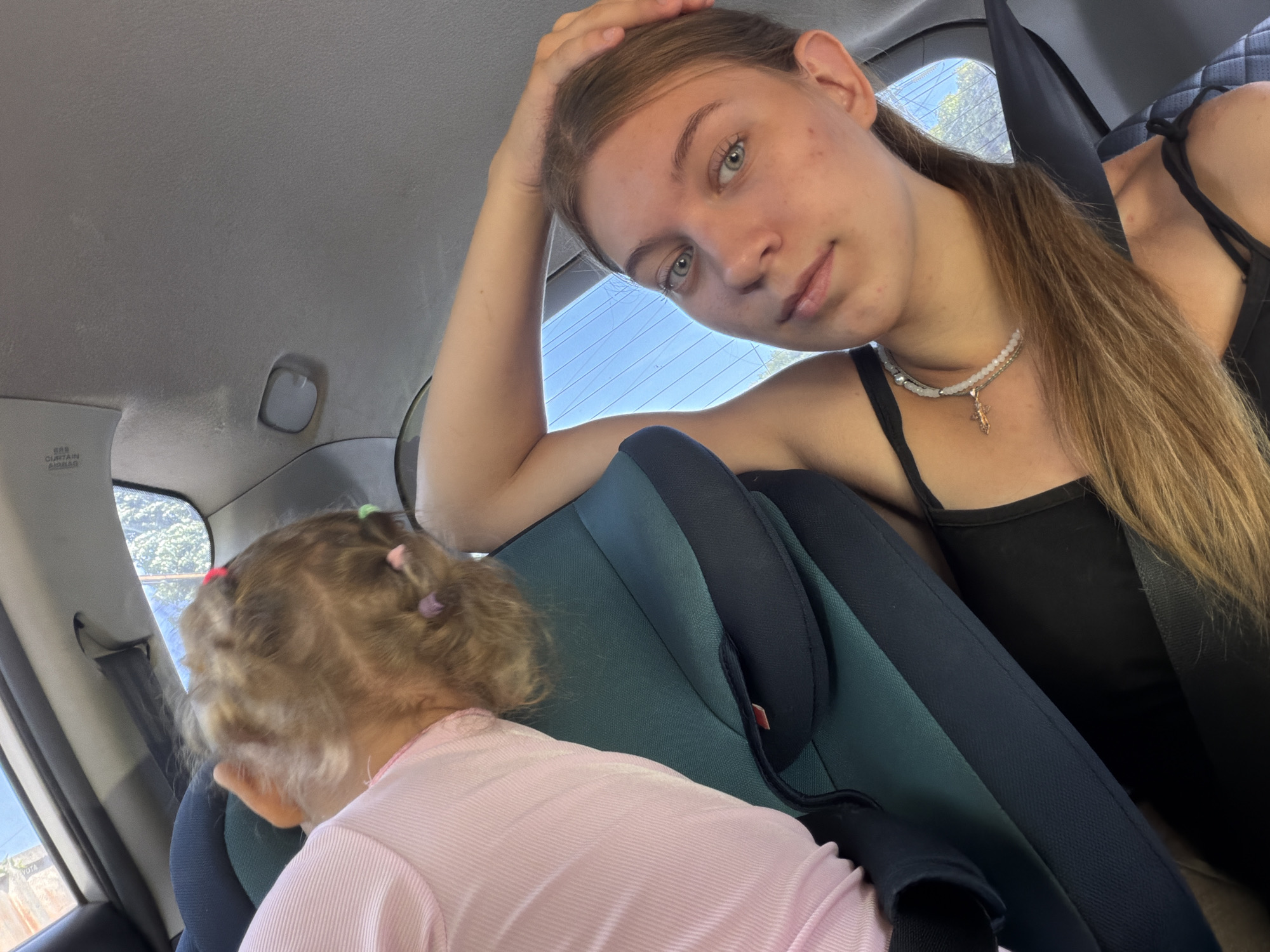 The image shows two individuals in the interior of a vehicle, likely a car. The individual in the foreground is a young woman with light brown hair, wearing a black tank top and a pearl necklace. She is looking directly at the camera with her right hand resting on her head. The second individual, a young child, is seated in a car seat in the background, facing away from the camera. The child has blonde hair and is wearing a pink top. The interior of the car appears to be grey and green, with a visible seatbelt. The view outside the car window suggests a sunny day with trees visible. The image appears to be a selfie taken by the young woman. The location of Rostov-on-Don, Russia, is not discernible from the visual information present in the image.