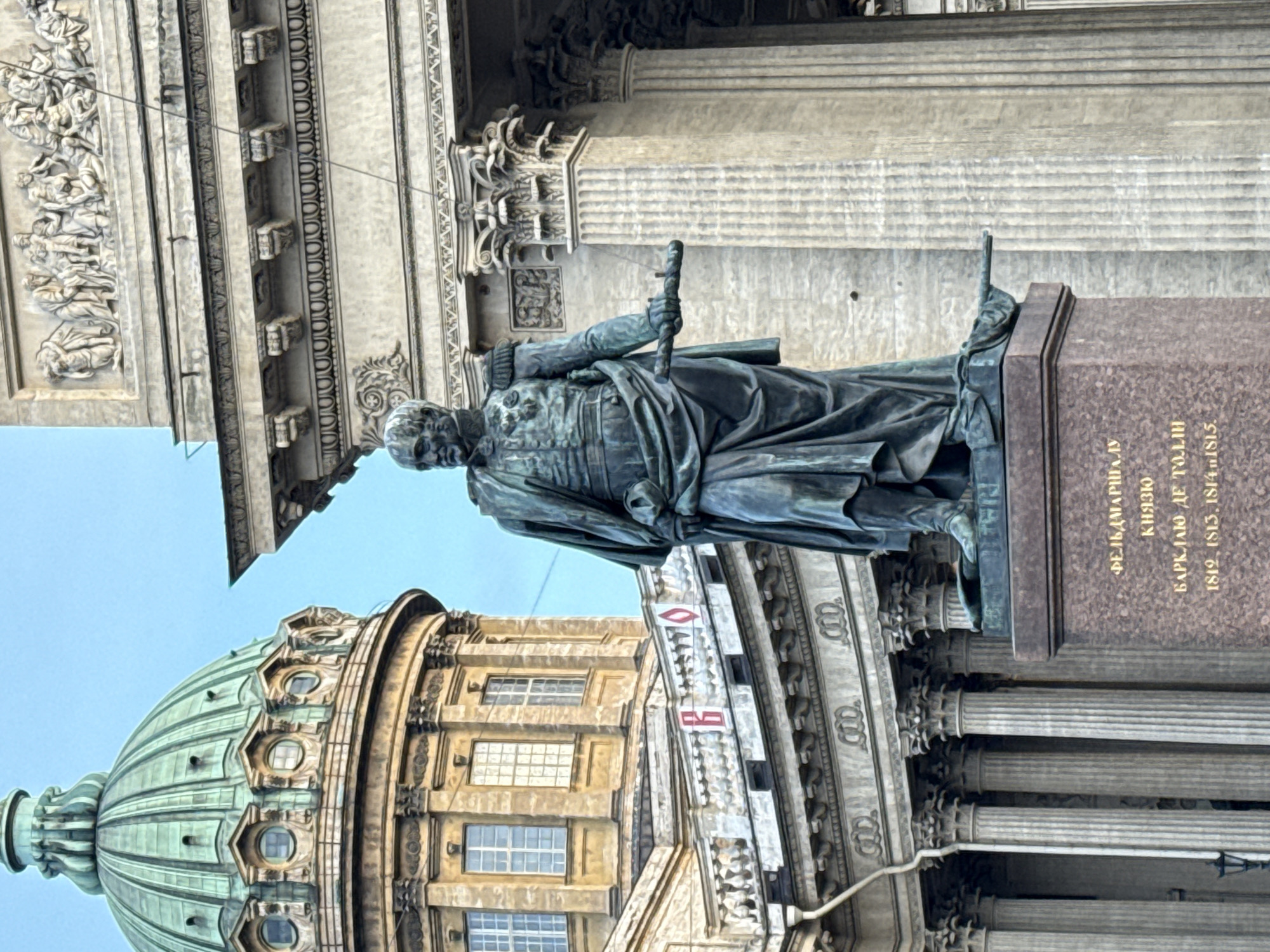 The image shows the monument to Field Marshal Barclay de Tolly in Saint Petersburg, Russia. The bronze statue stands on a granite pedestal. The statue depicts a man in military uniform holding a baton. In the background is the architectural facade of a building with columns and a dome. The sky is visible above the buildings.